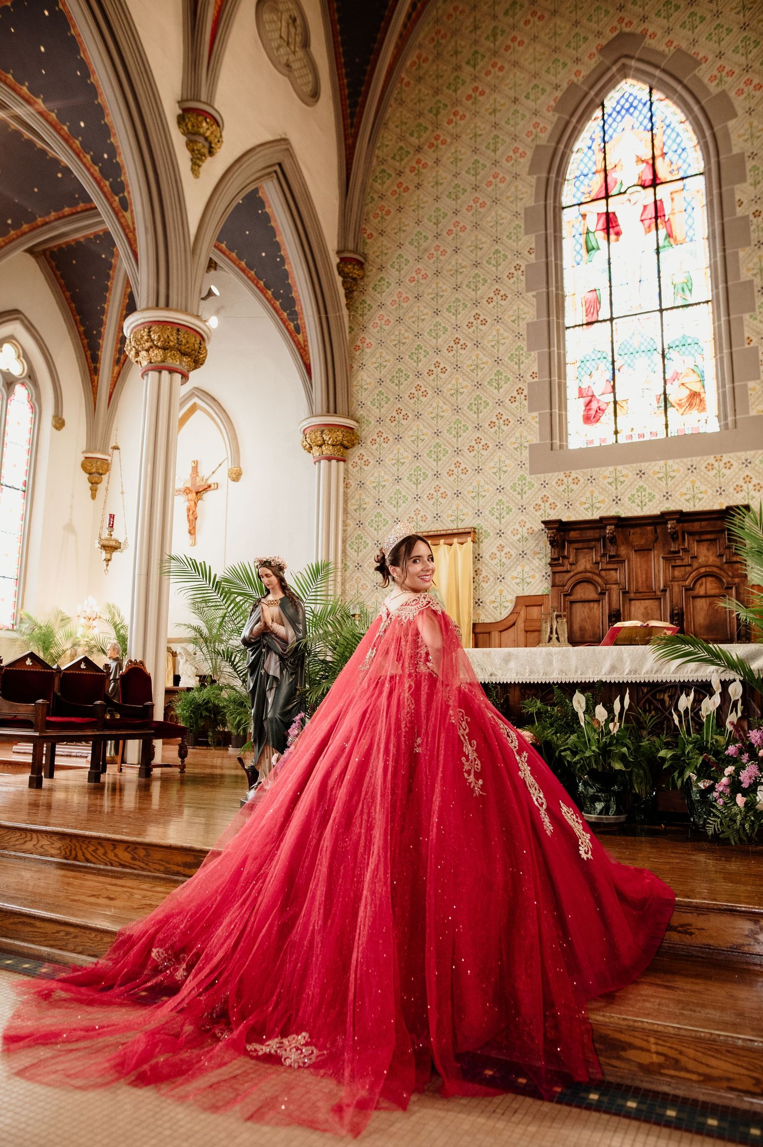 Quinceañera in flowing red tulle ball gown twirling near church altar with stained glass windows.