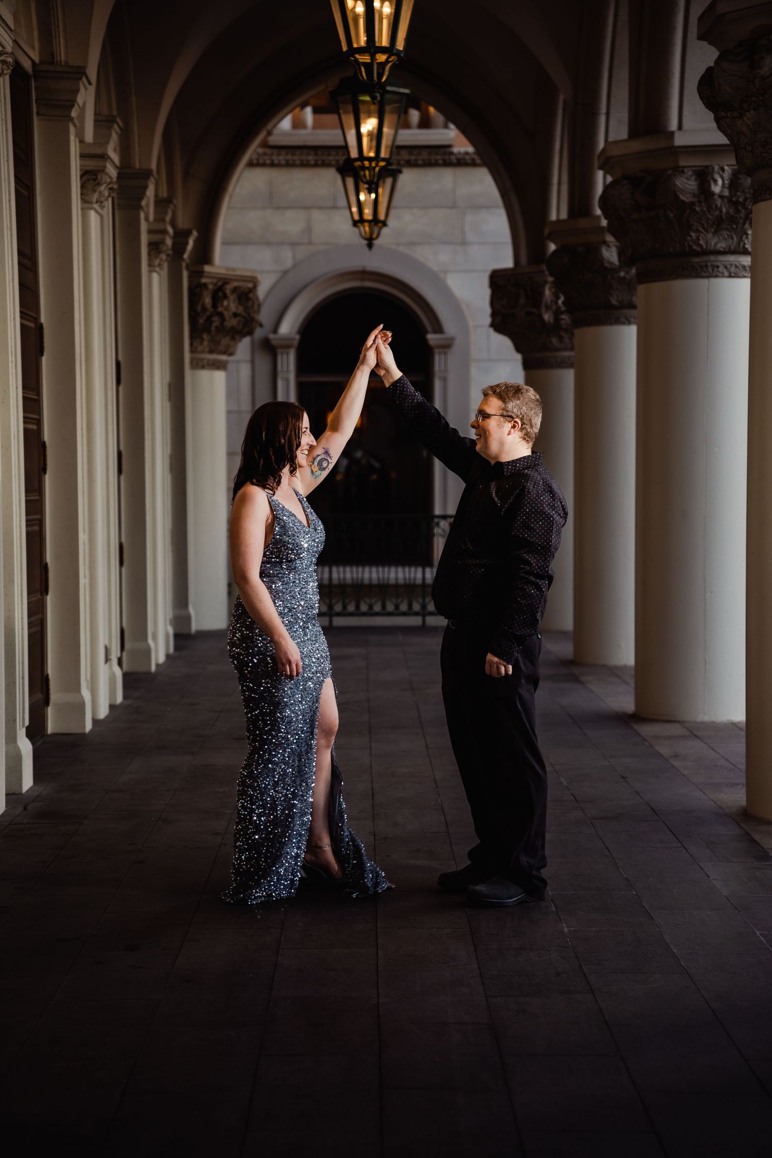 Couple dancing together under elegant arched columns at night, woman in silver sequin gown, man in black.