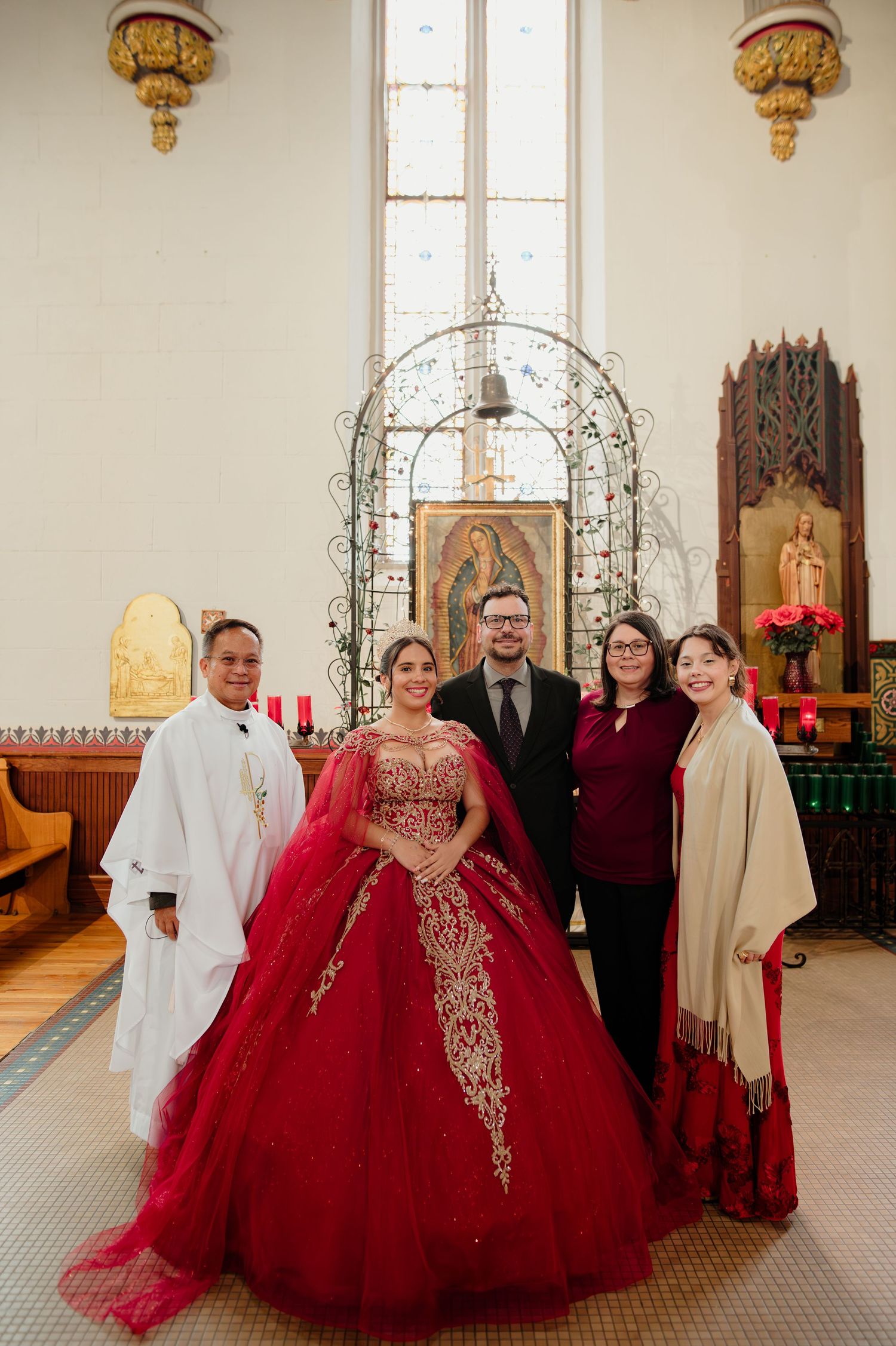 Quinceañera in red gown poses with priest and family members before a Virgin of Guadalupe shrine.