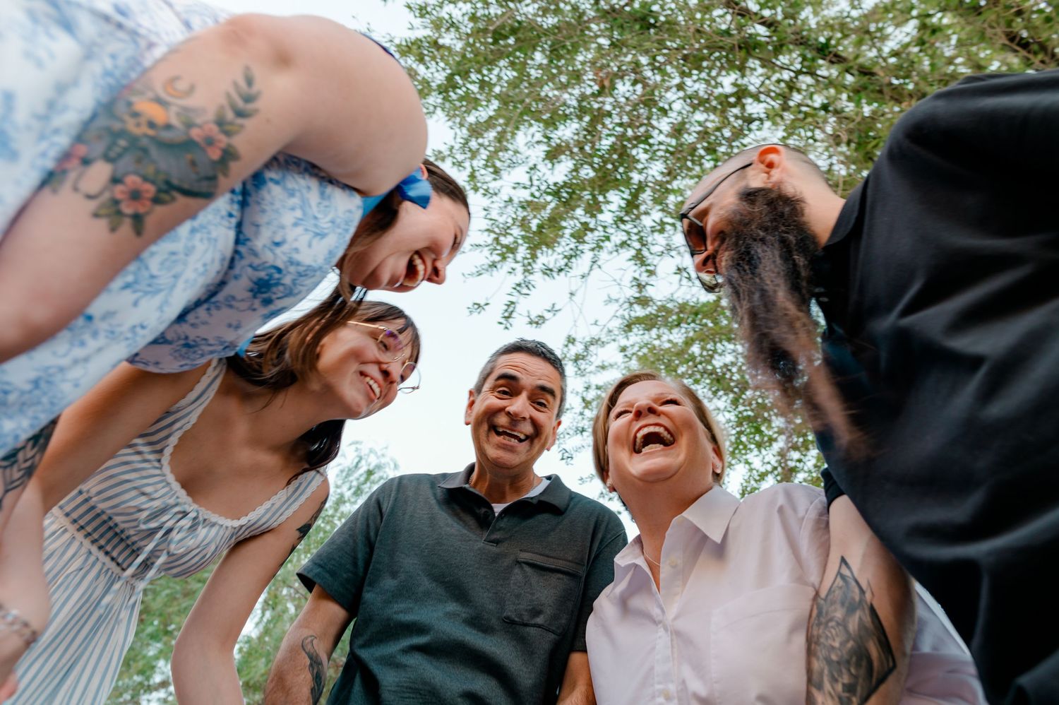 Happy family of five huddles together laughing in low-angle shot beneath green tree canopy.