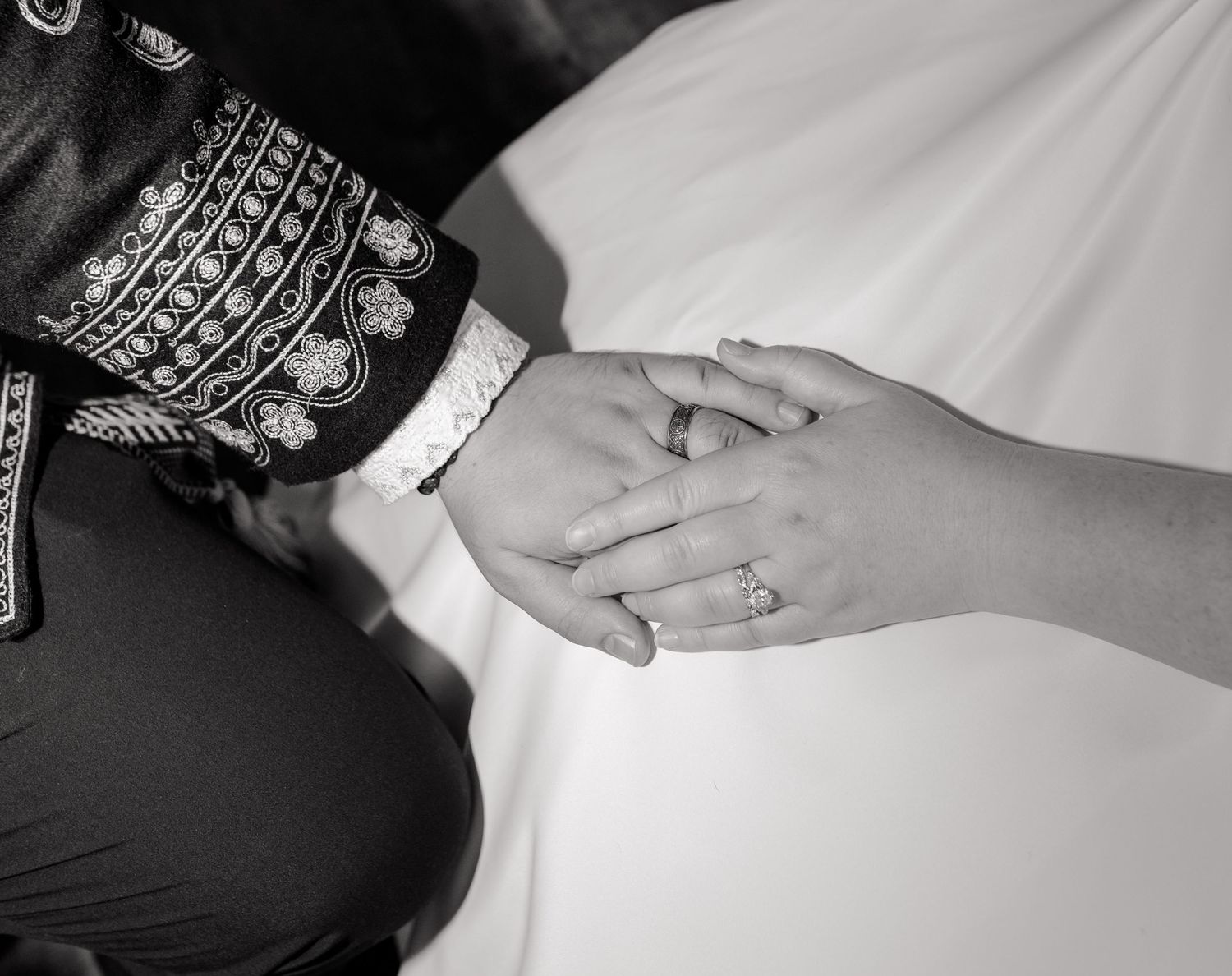 Black and white photo of a bride and groom holding hands, showing wedding rings, in traditional attire.