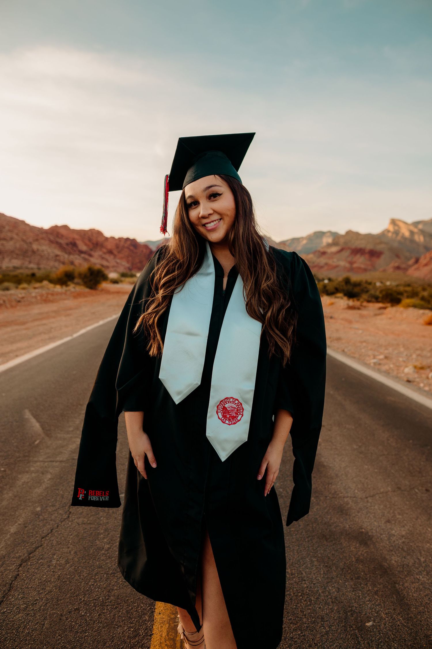 Smiling graduate in black cap and gown with stole poses on desert road at sunset.