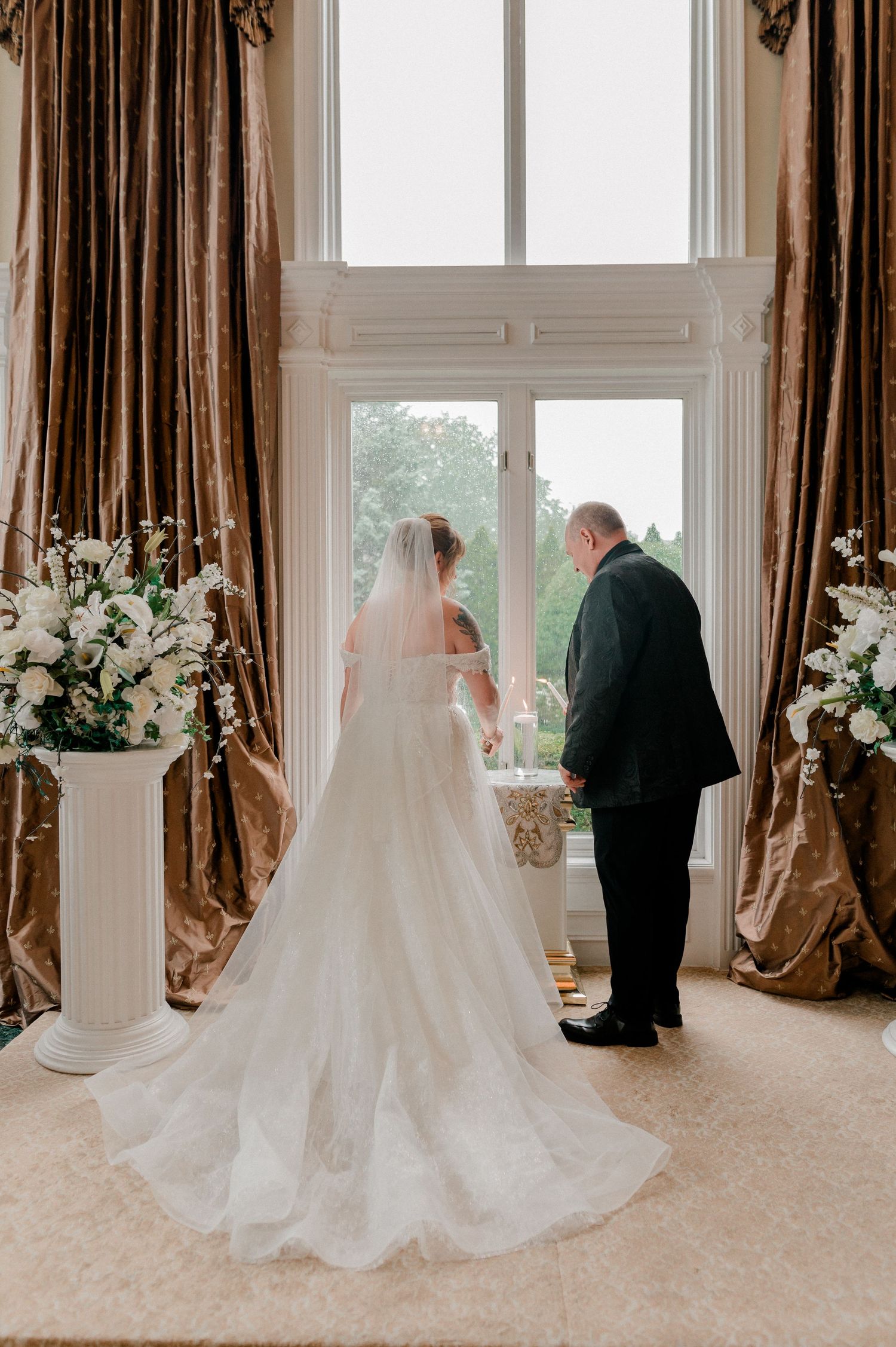 Bride in white gown and veil stands with groom before large window flanked by floral arrangements and gold drapes.