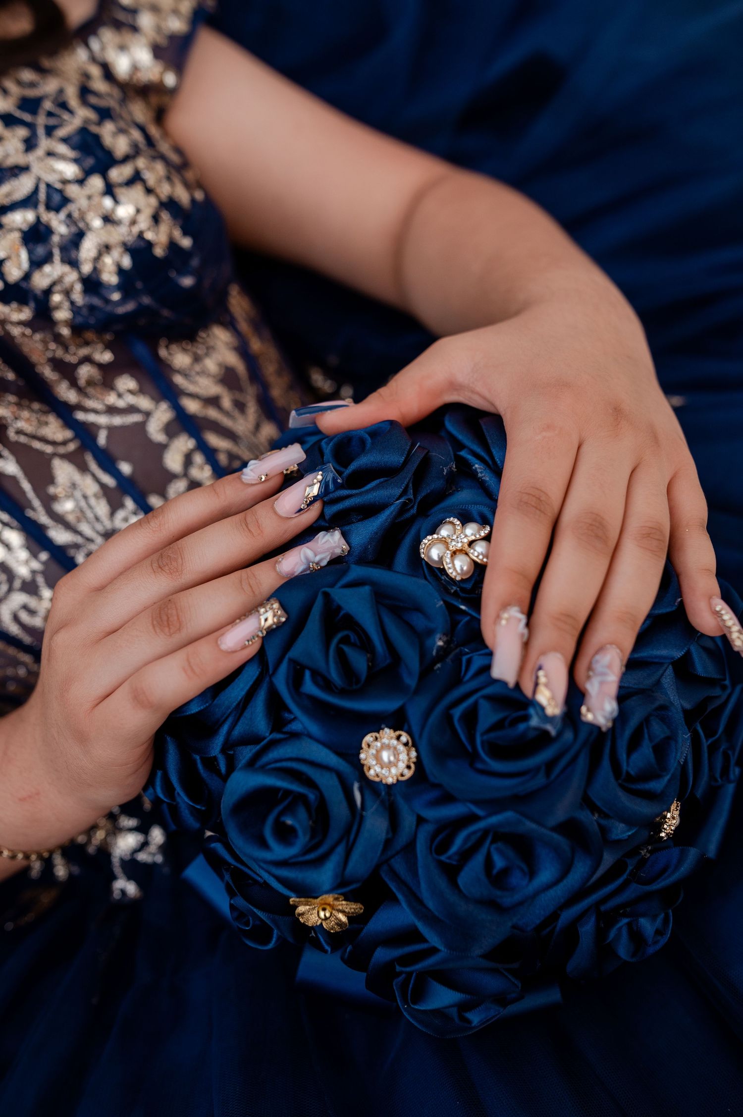 Close-up of elegant blue satin rose bouquet with pearl brooches held by hands with ornate acrylic nails in navy gown.