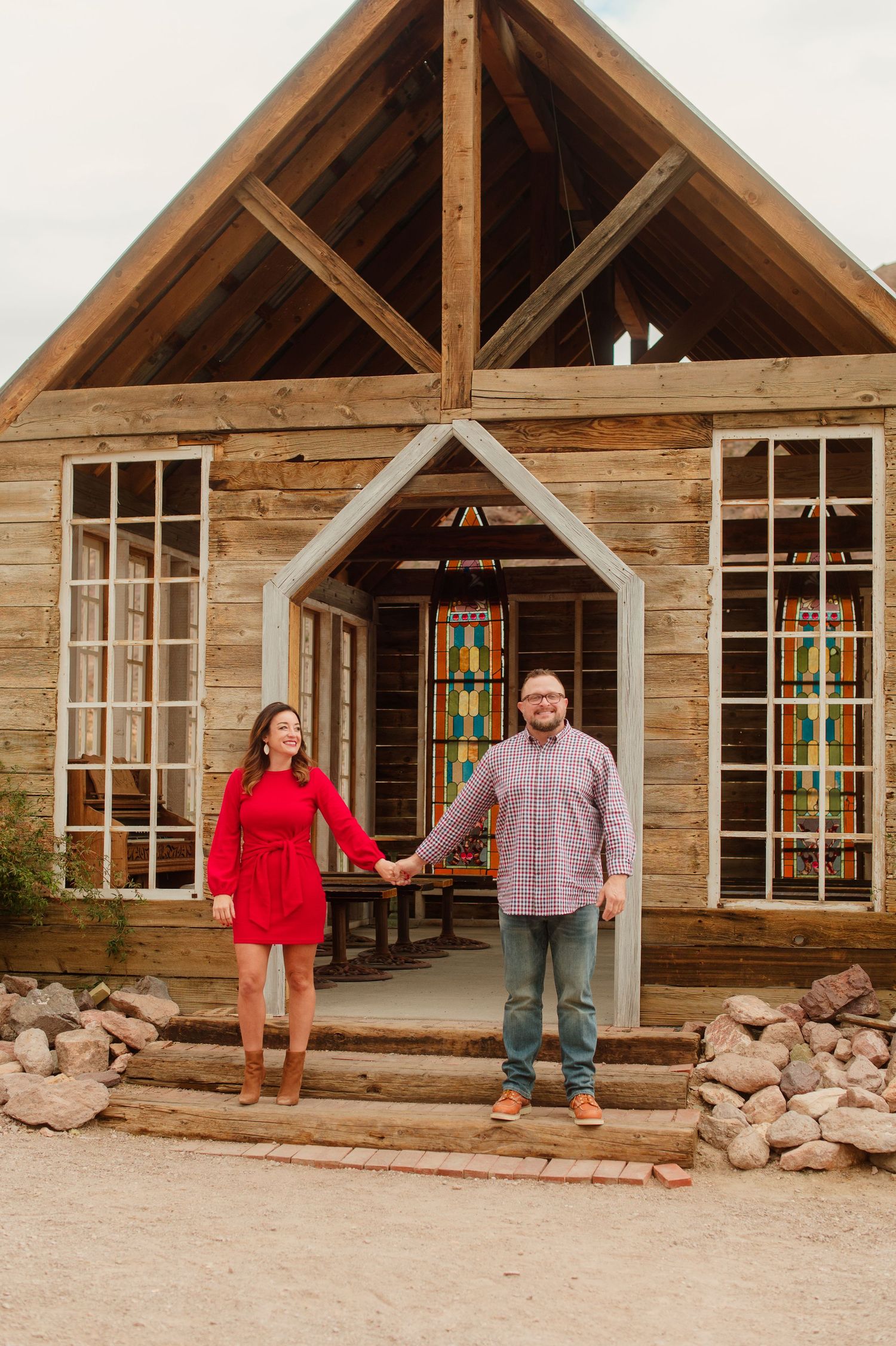 Couple holding hands in front of a wooden chapel with stained glass windows outdoors.