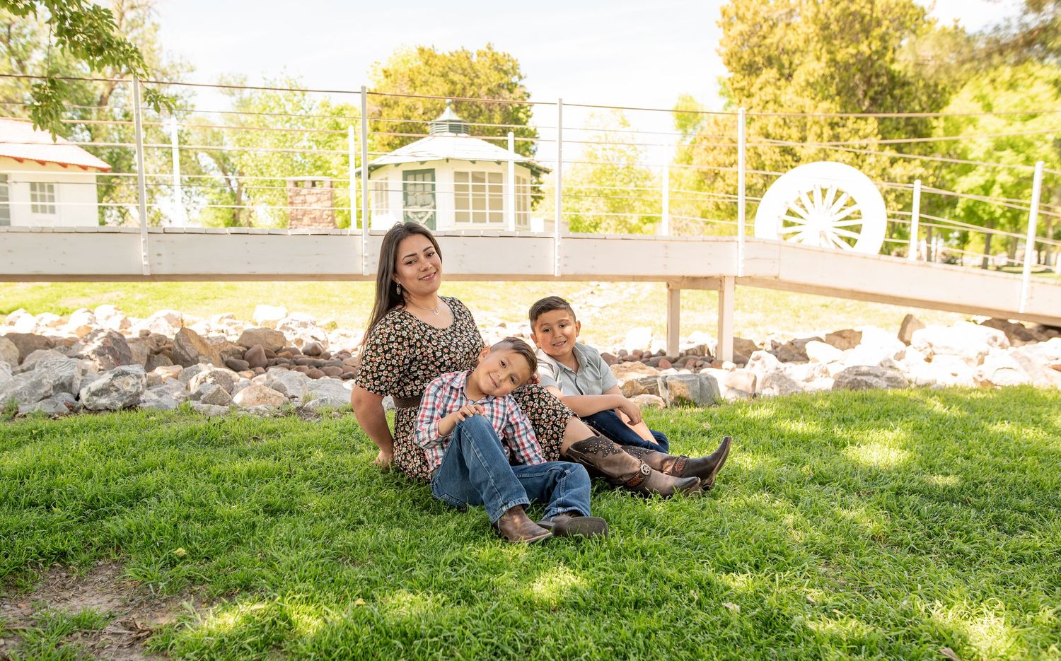 Mother sitting on grass with two young sons in a sunny park setting.