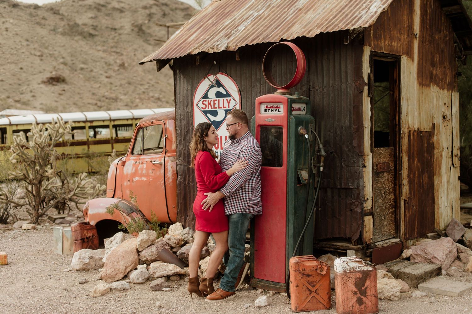 Couple embracing near a retro red gas pump outside a weathered tin-roofed desert shack.