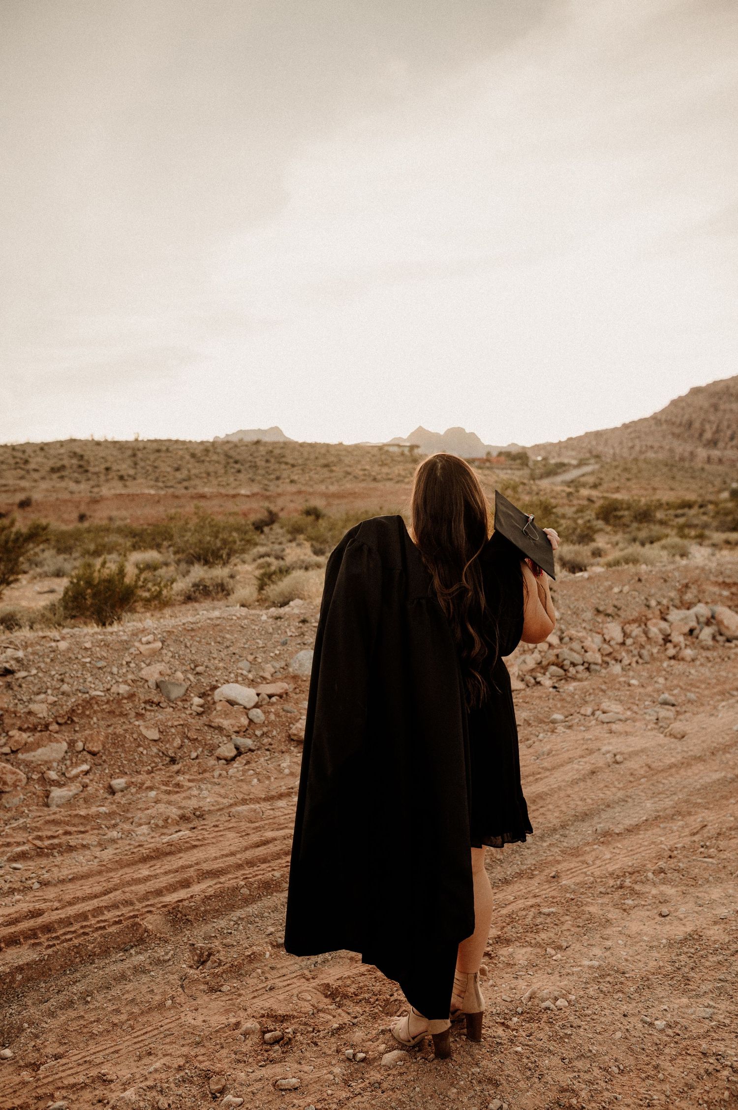 Graduate in black gown walks away on rocky desert path holding mortarboard cap.
