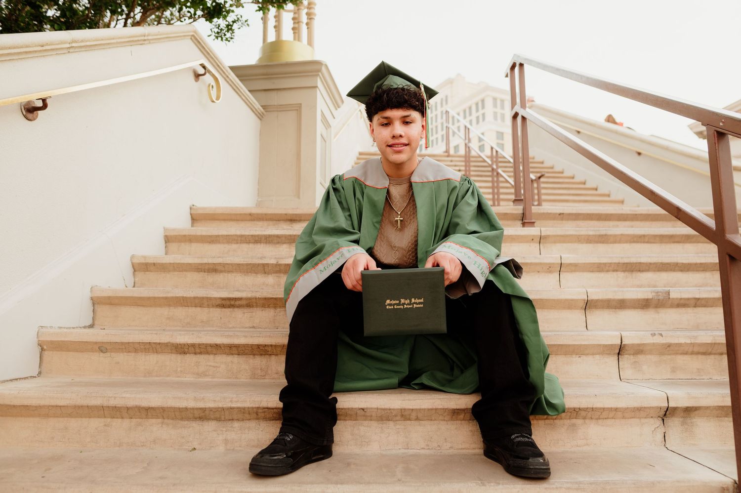 Graduate in green cap and gown sitting on steps holding diploma at Caesars Palace.