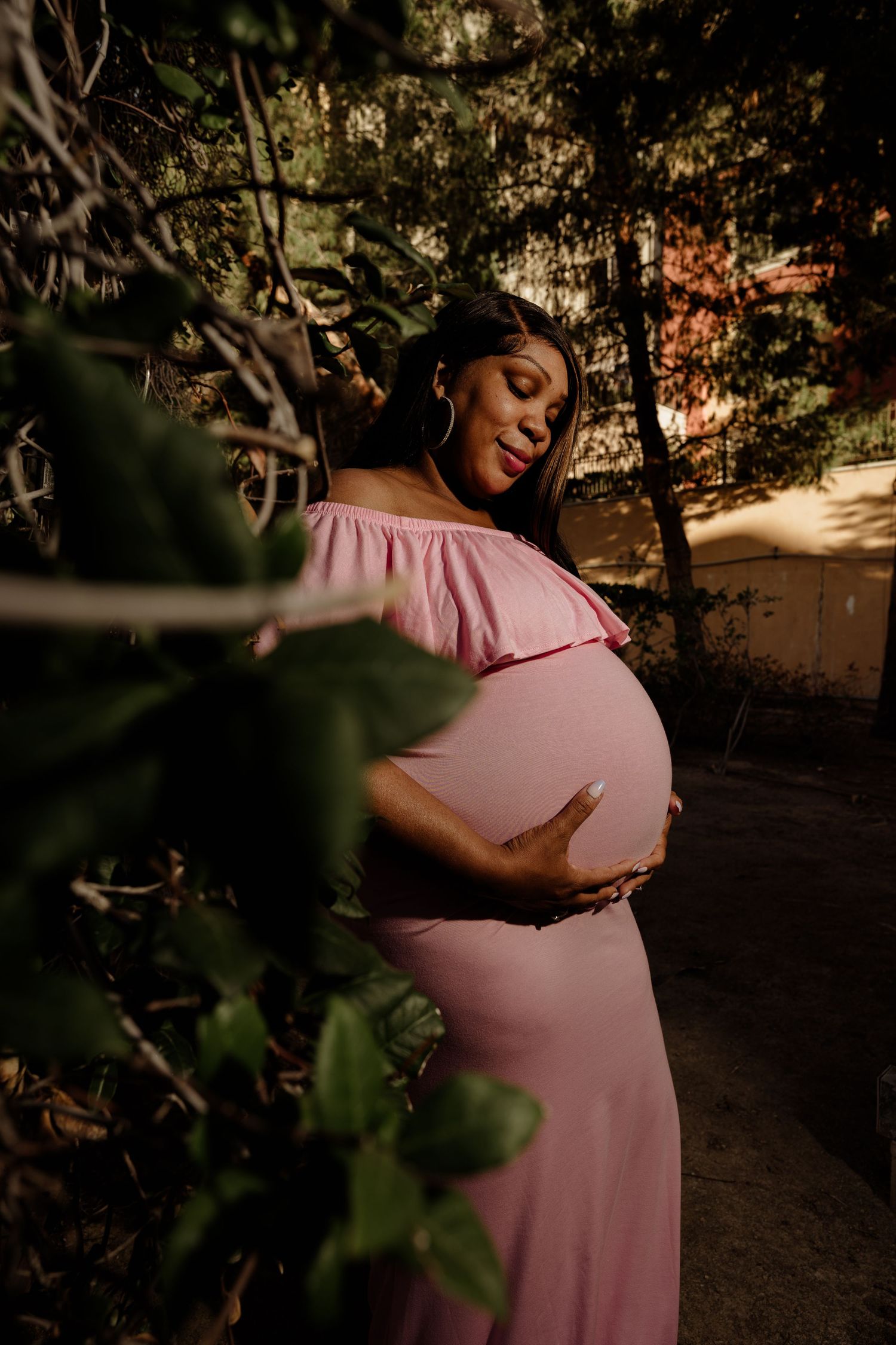 Pregnant woman in pink off-shoulder maternity gown cradling her baby bump among lush green trees.