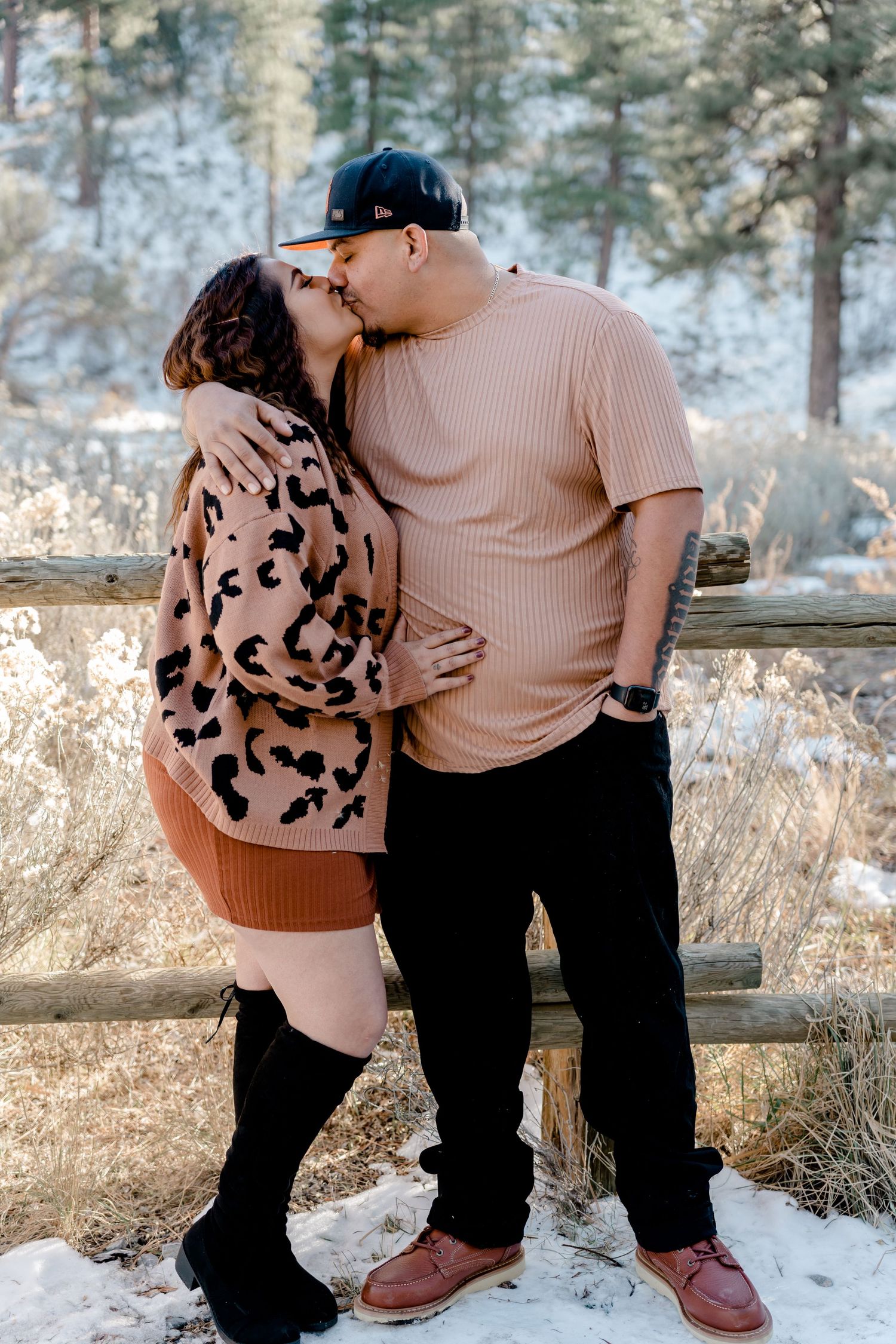 A couple shares a romantic kiss outdoors in a snowy winter setting near a rustic wooden fence.