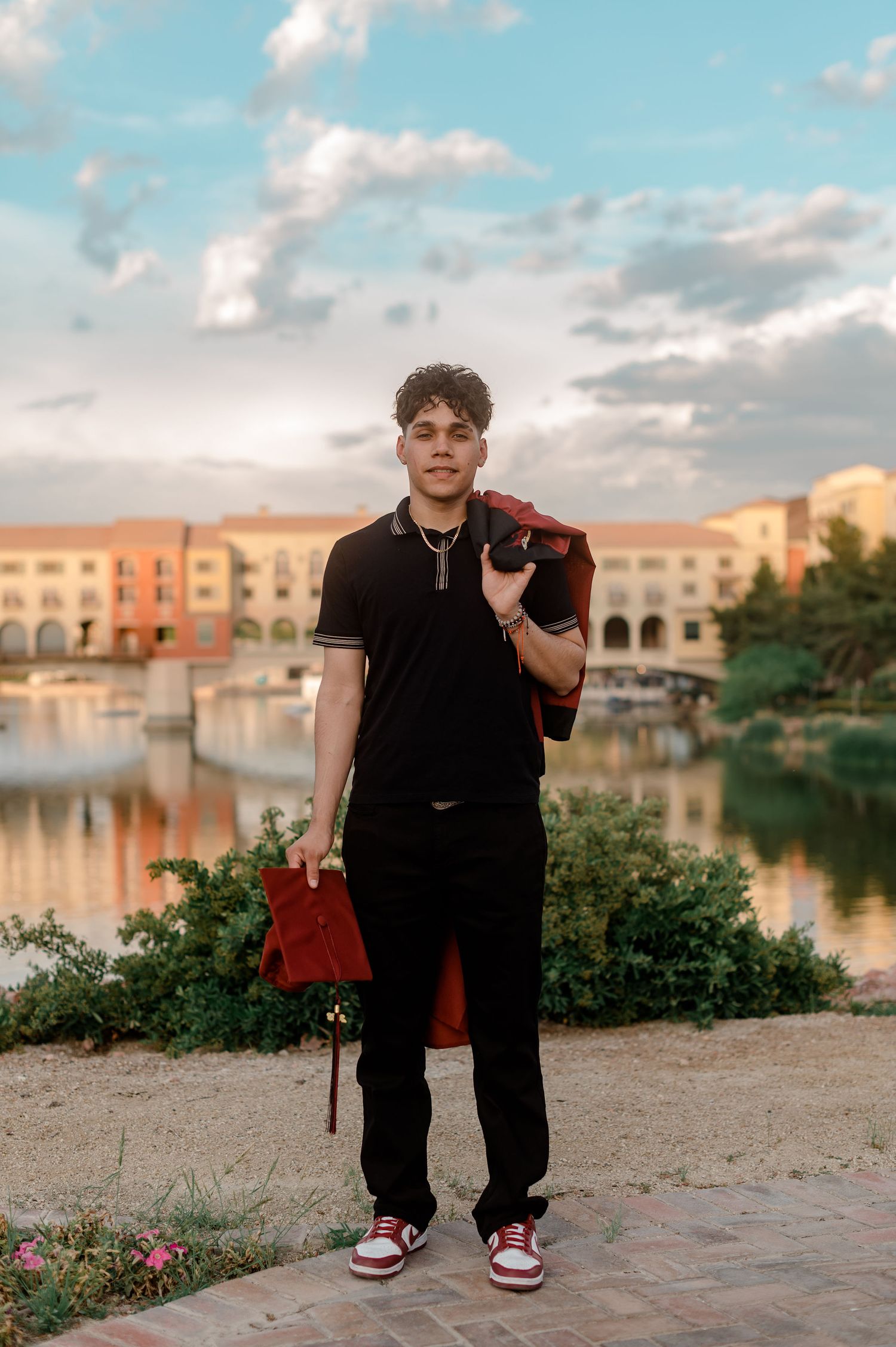 Graduate holding red cap and gown by scenic waterfront at golden hour.