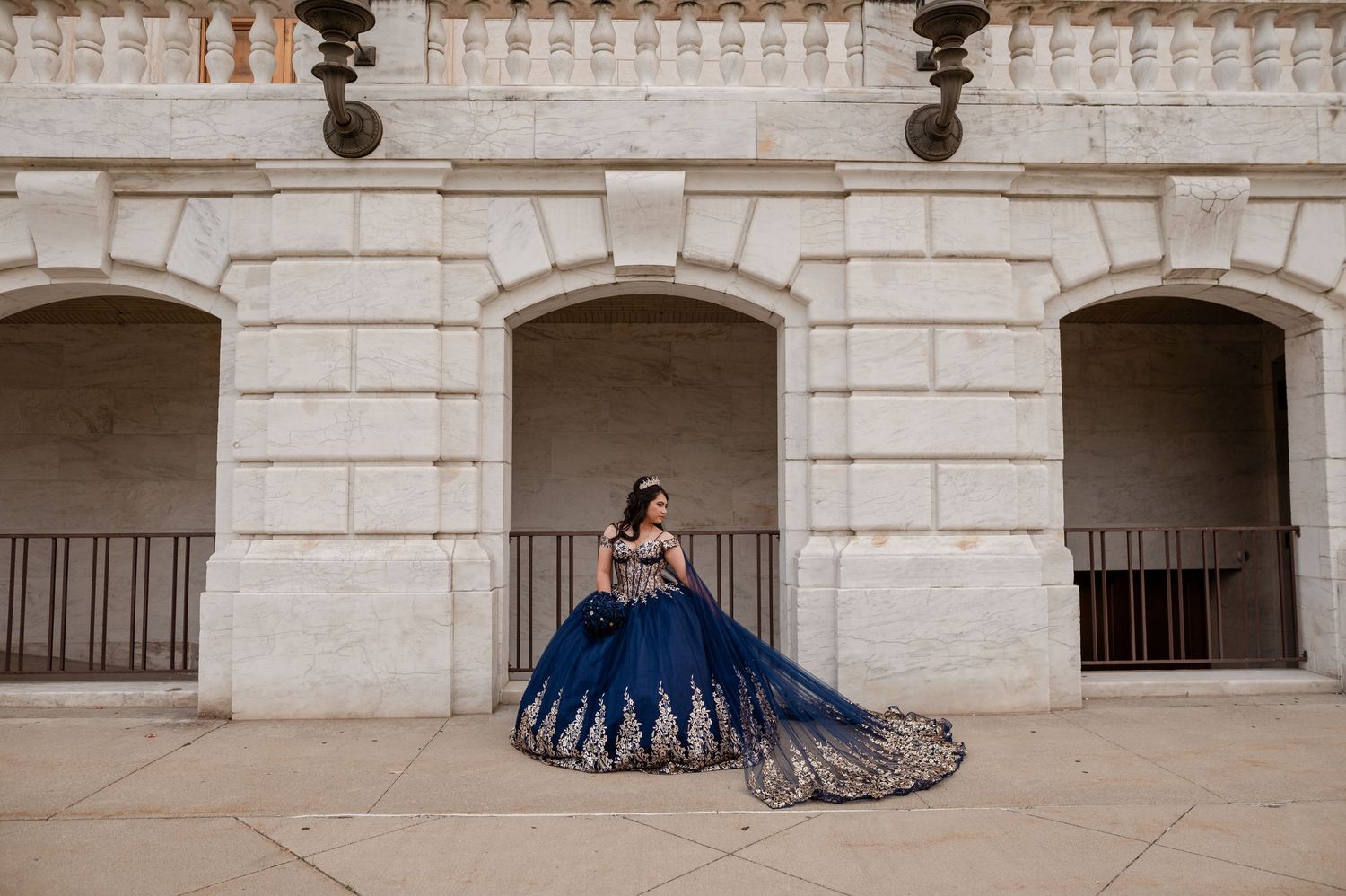 A young woman in a navy blue and gold quinceañera ball gown poses elegantly in front of stone arched walls.
