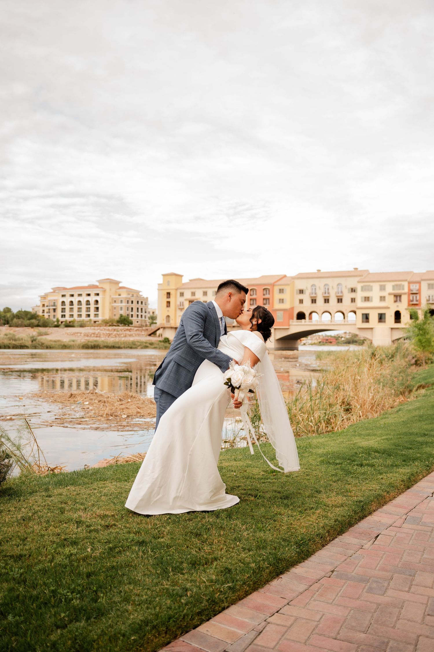 Groom dips bride for a romantic kiss beside a scenic waterway with a bridge backdrop.
