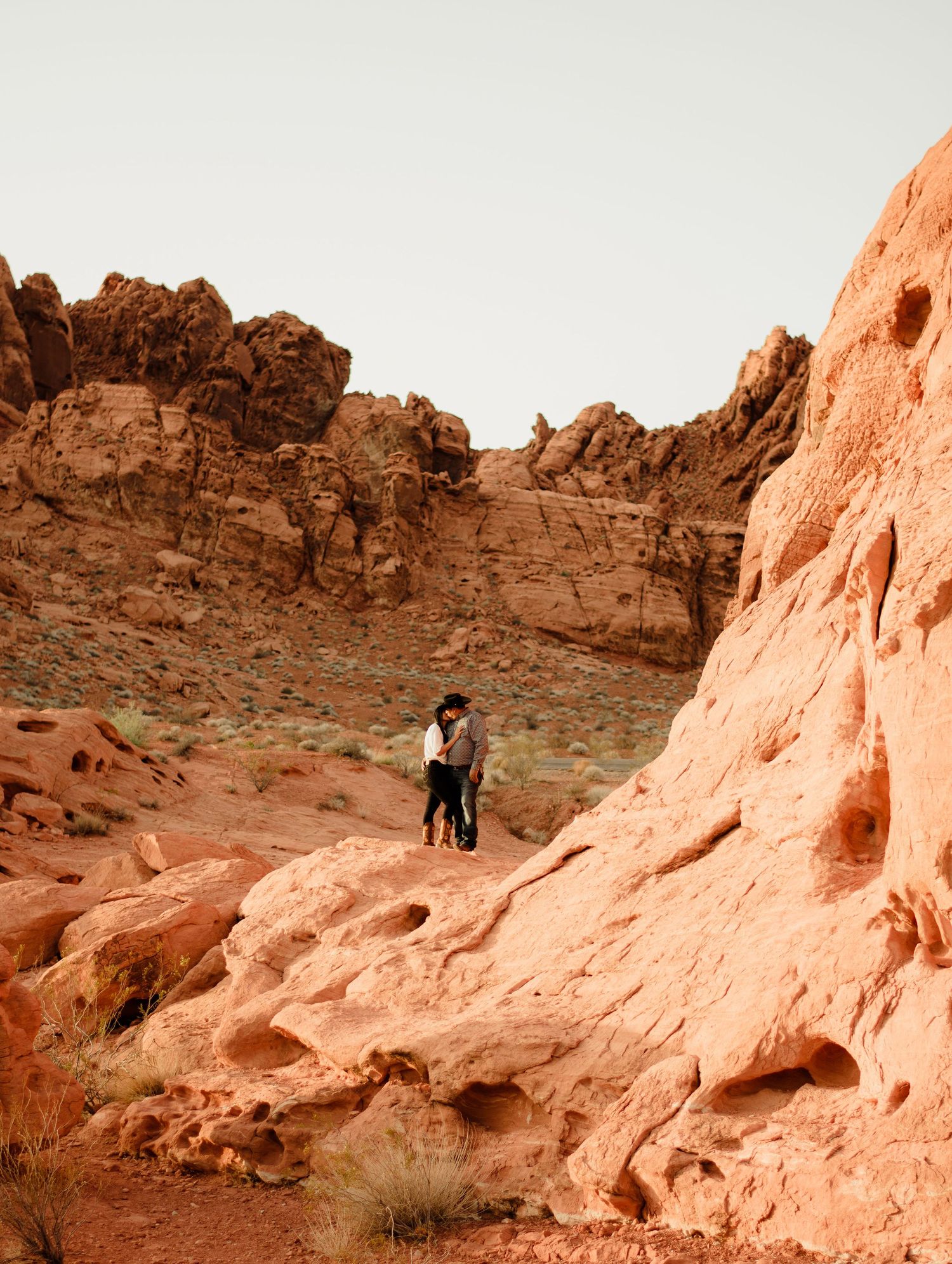 Couple embracing on red rock terrain surrounded by dramatic desert canyon landscape.