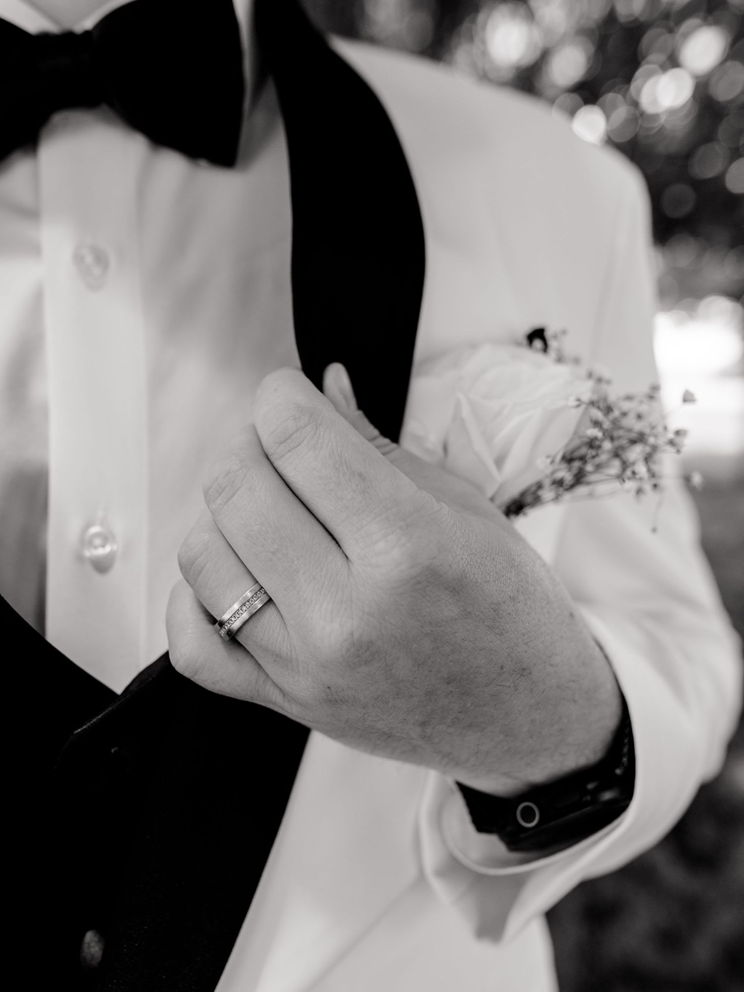Black and white photo of groom in white tuxedo with bow tie, wearing wedding band, holding bride close at wedding.