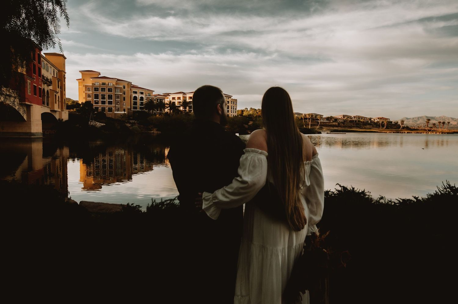 Bride and groom embrace while gazing at a scenic waterfront view with colorful buildings reflected in the water.