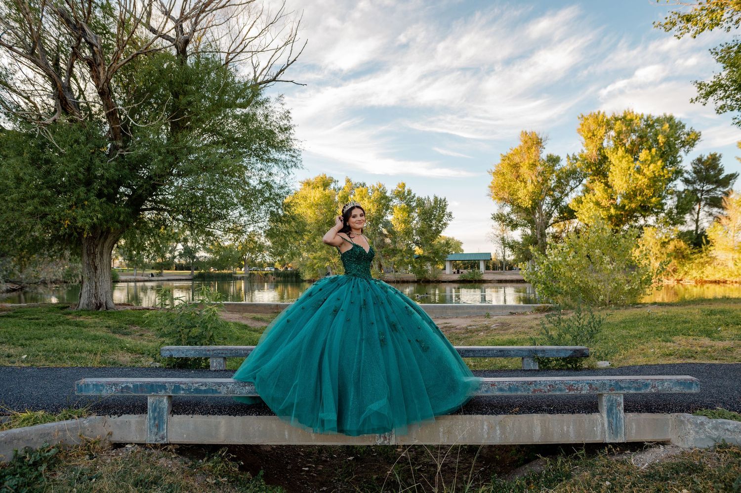 Woman in full emerald green quinceañera ball gown stands on a bridge surrounded by autumn trees.