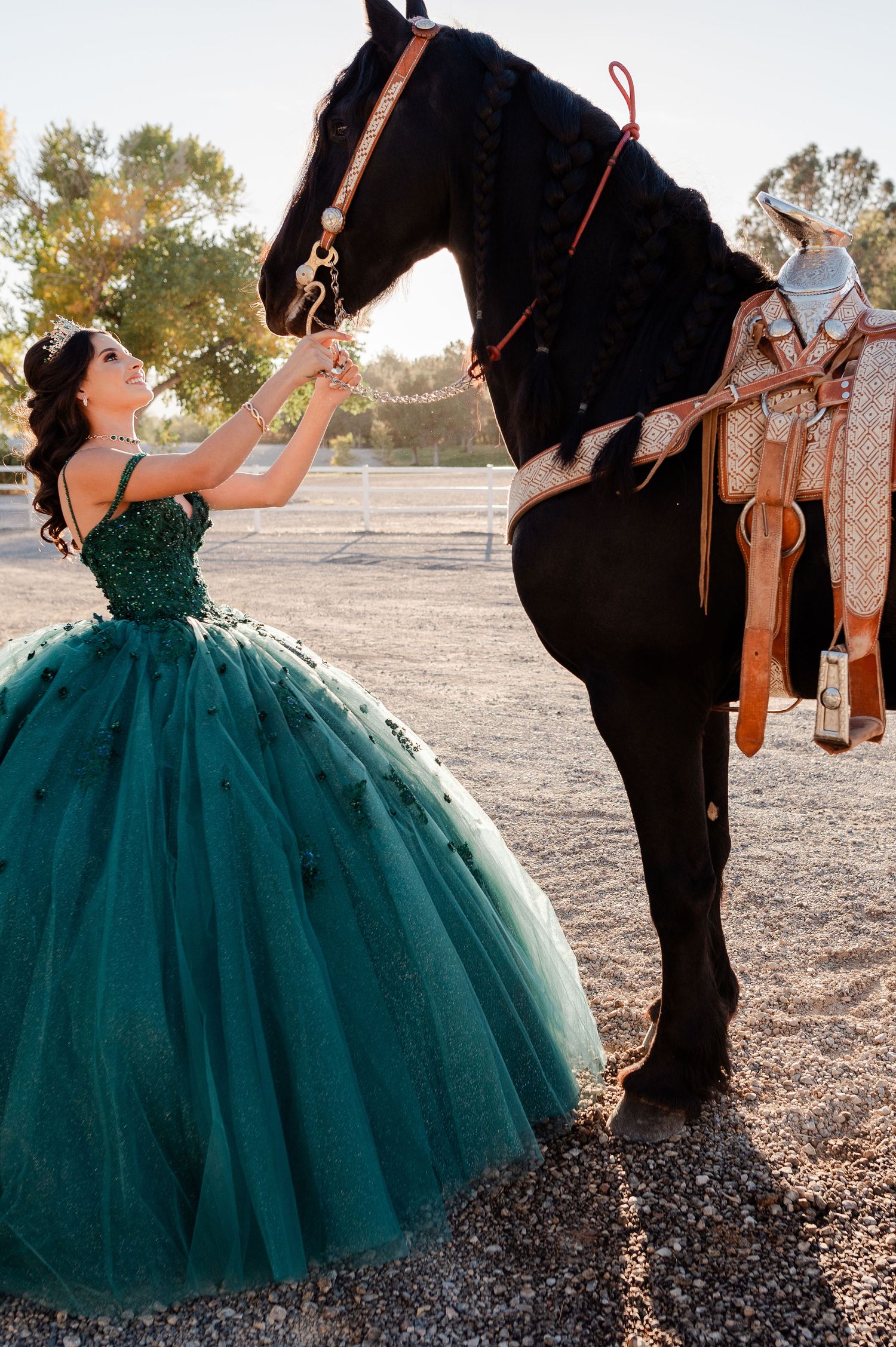 Quinceañera girl in green tulle ball gown reaching up to a saddled black horse outdoors.
