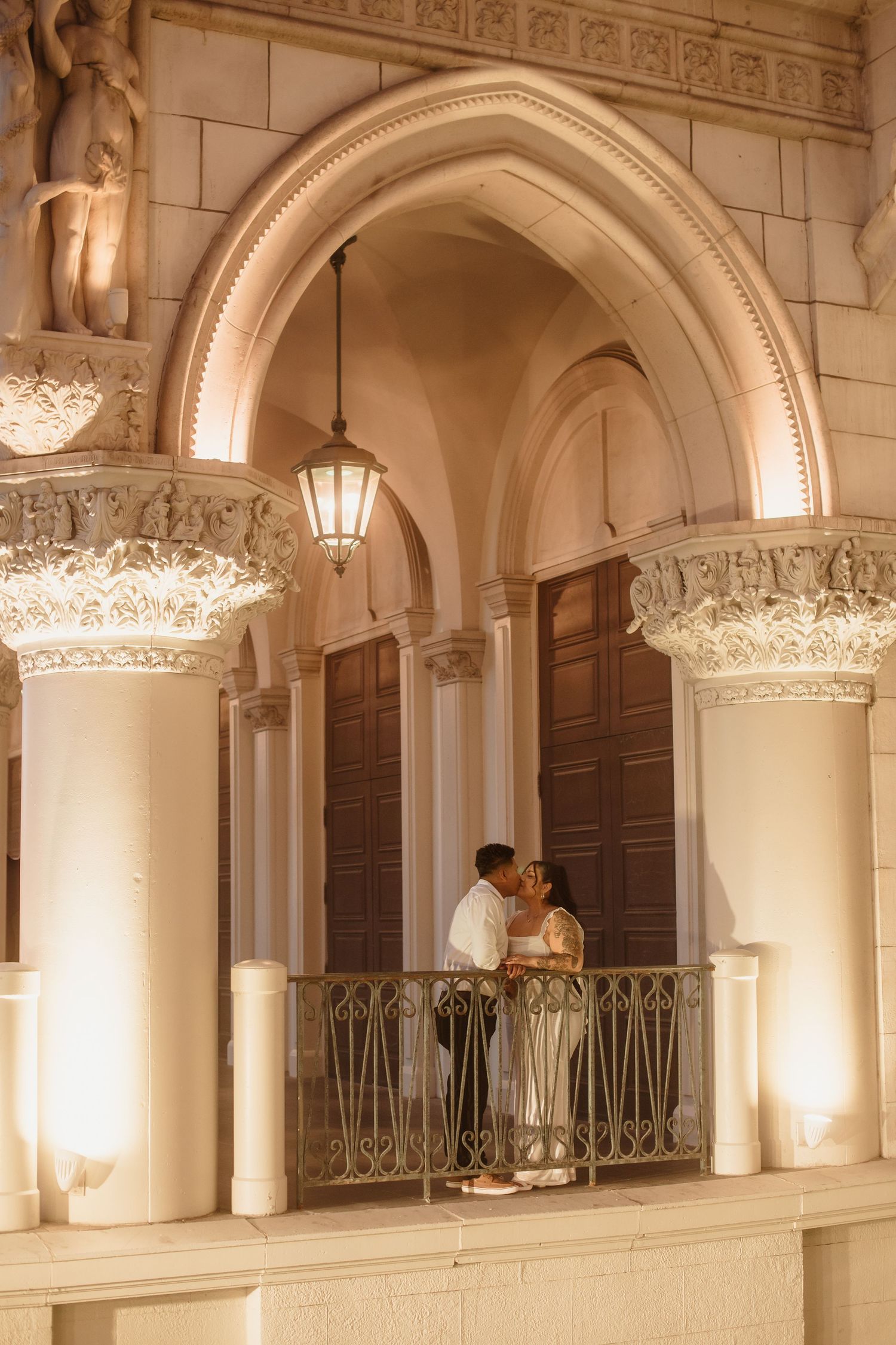 Couple sharing a romantic kiss on an ornate balcony beneath a grand illuminated arch with decorative columns at night.