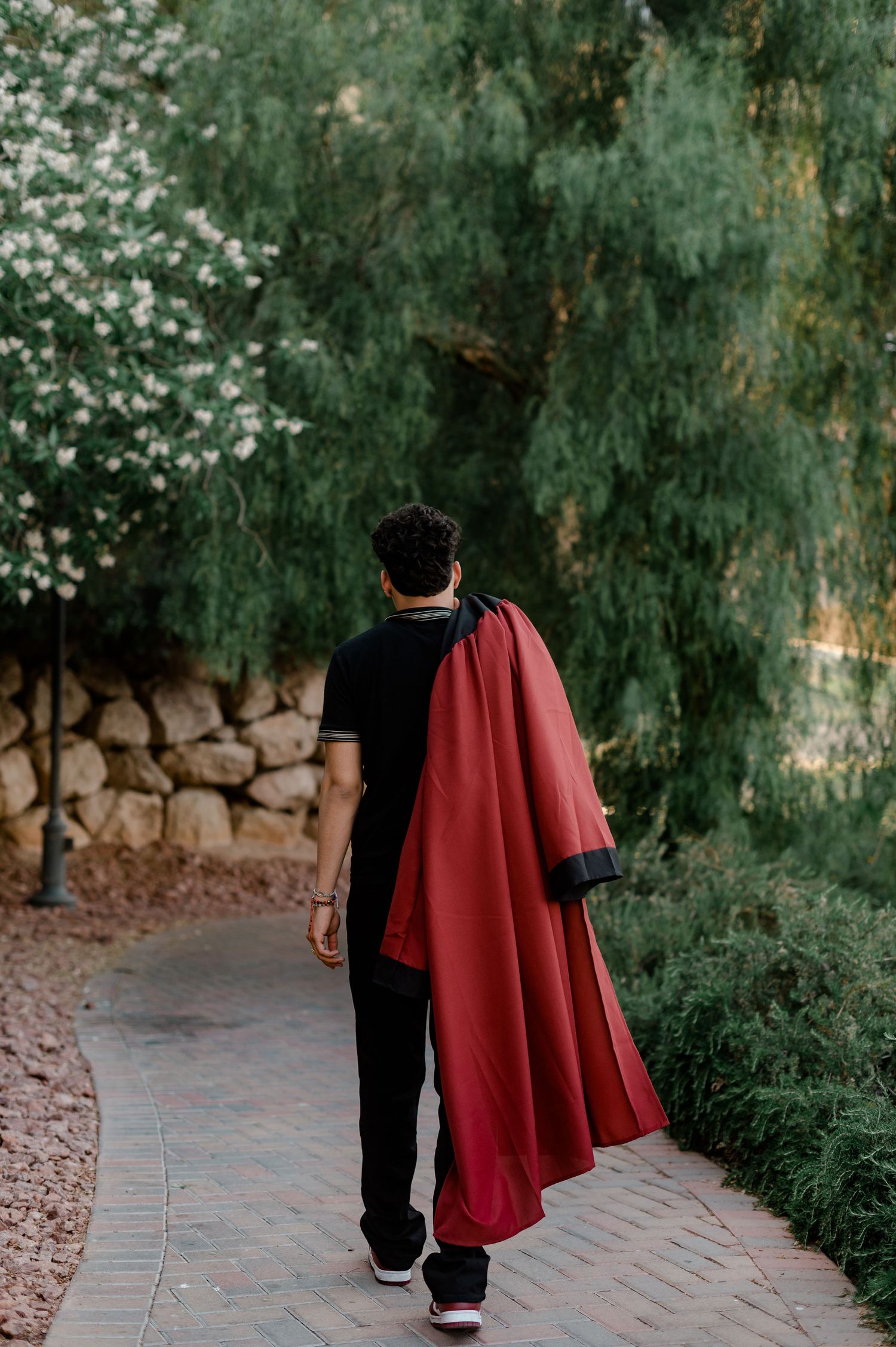 Graduate walks away on brick path with red gown draped over shoulder near trees.