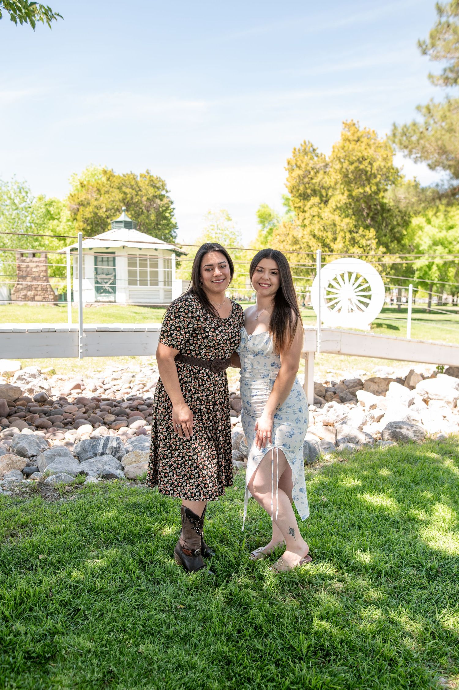 Two women in floral dresses pose together in a sunny park with a gazebo and wagon wheel background.