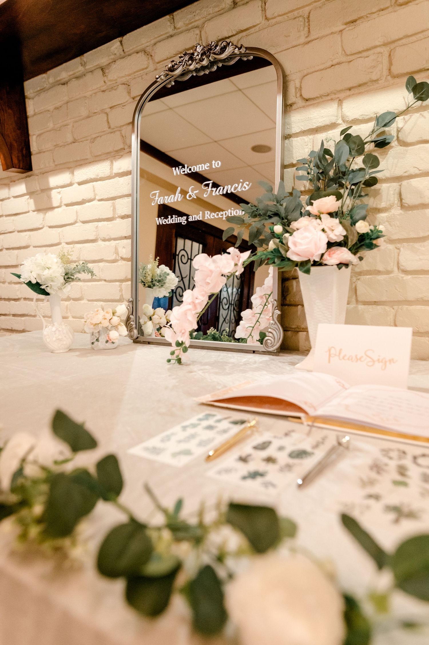Elegant wedding welcome mirror sign with floral arrangements, guest book, and white brick wall backdrop.