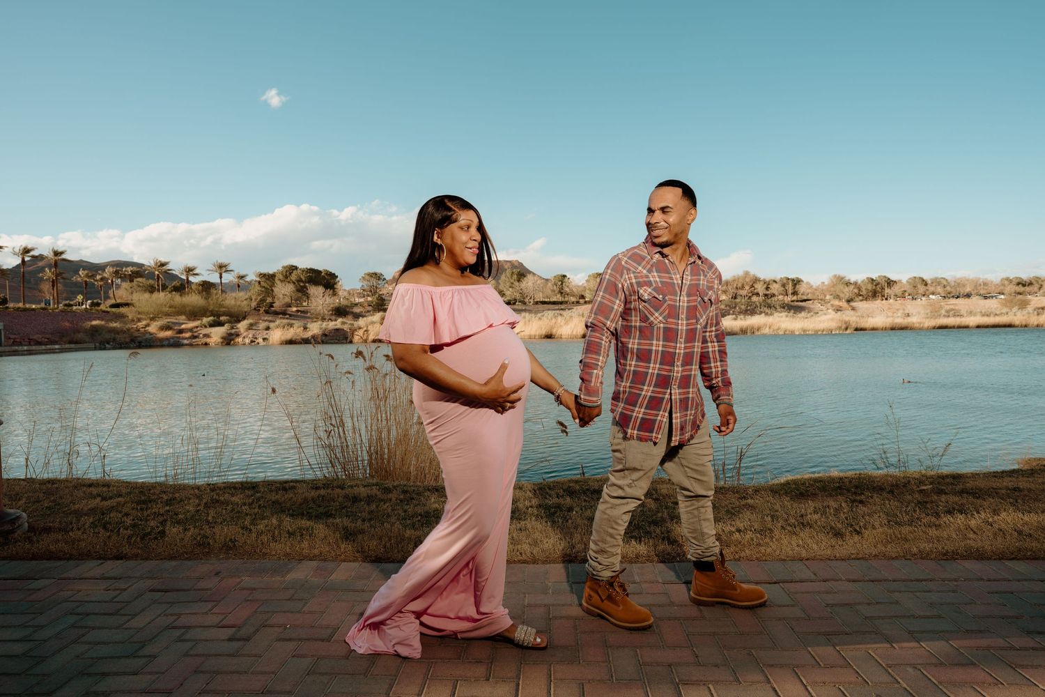 Pregnant woman in pink off-shoulder gown walks with partner along lakeside brick path under blue sky.