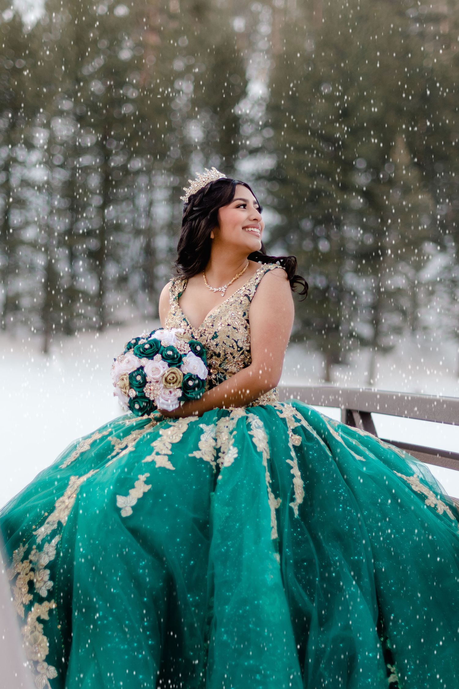 Young woman in emerald green and gold quinceañera gown holding bouquet in snowy winter wonderland setting.