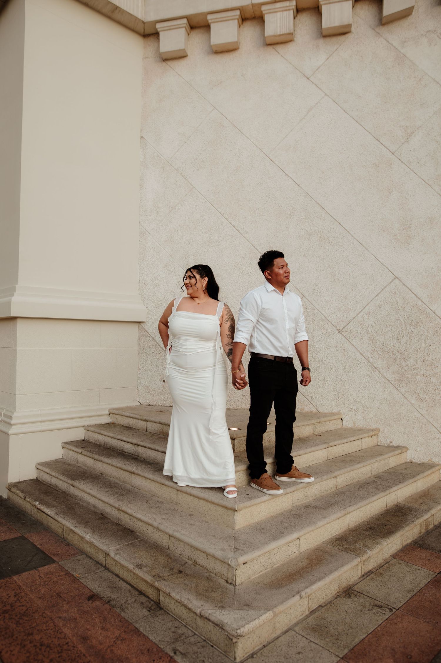 Couple in wedding attire hold hands while standing on steps of grand architectural building.