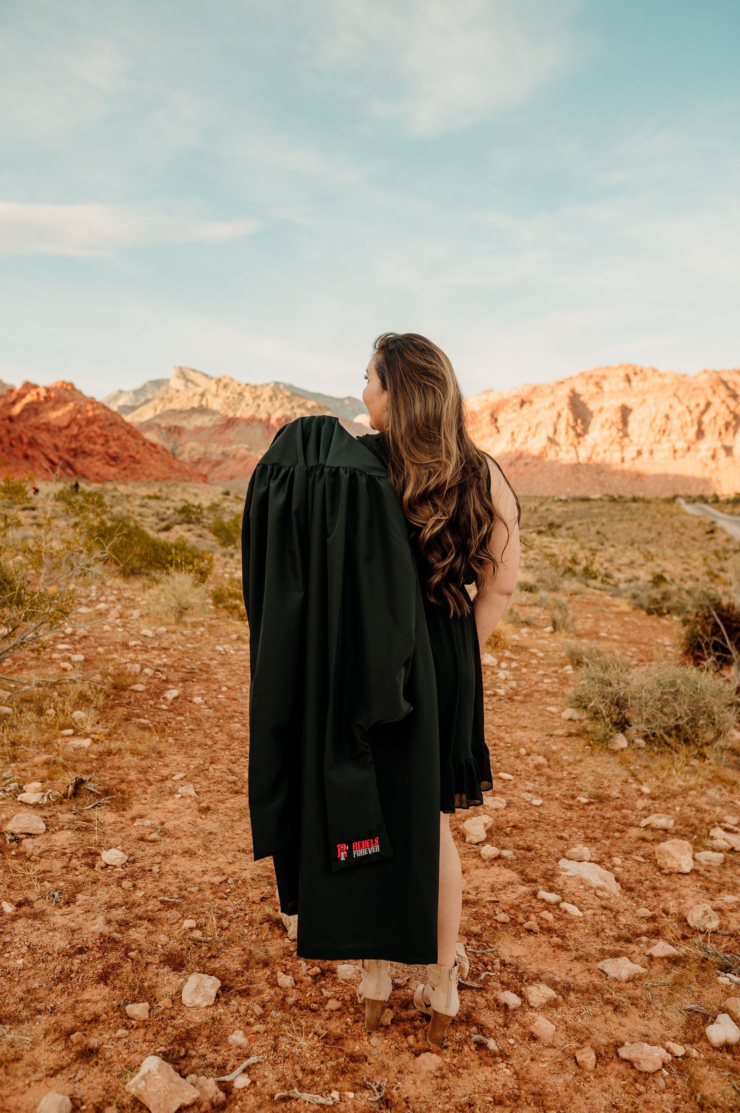 Woman in black graduation gown stands with back to camera overlooking Red Rock Canyon.