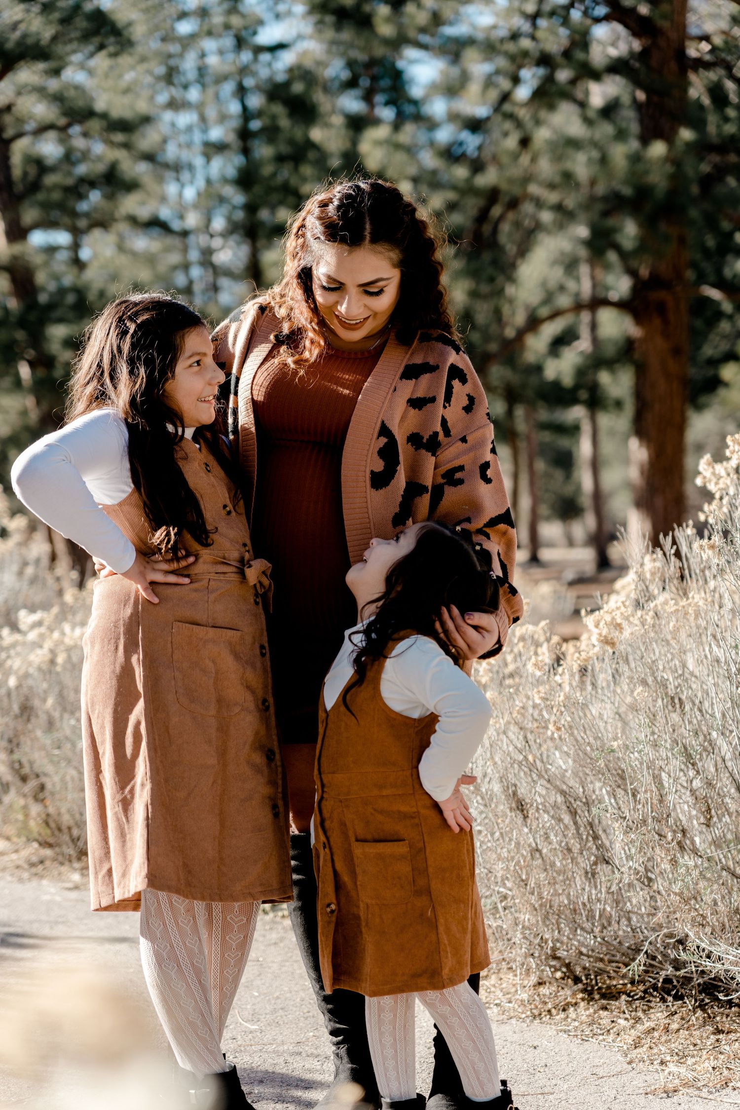 Mother and two daughters in matching brown outfits walking on sunlit forest path.