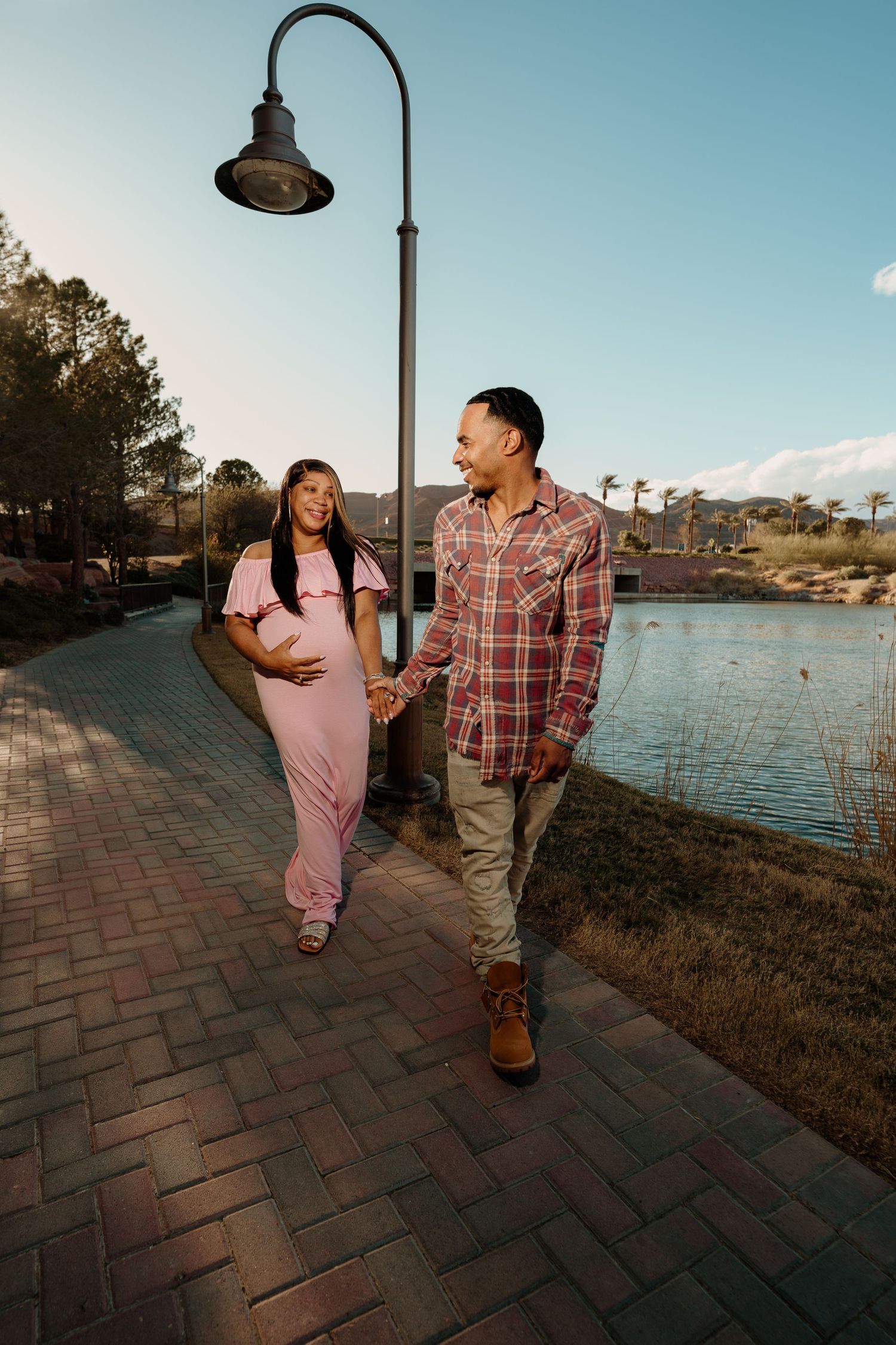 Expecting couple walks hand-in-hand on brick path by lake at sunset, woman in pink jumpsuit.