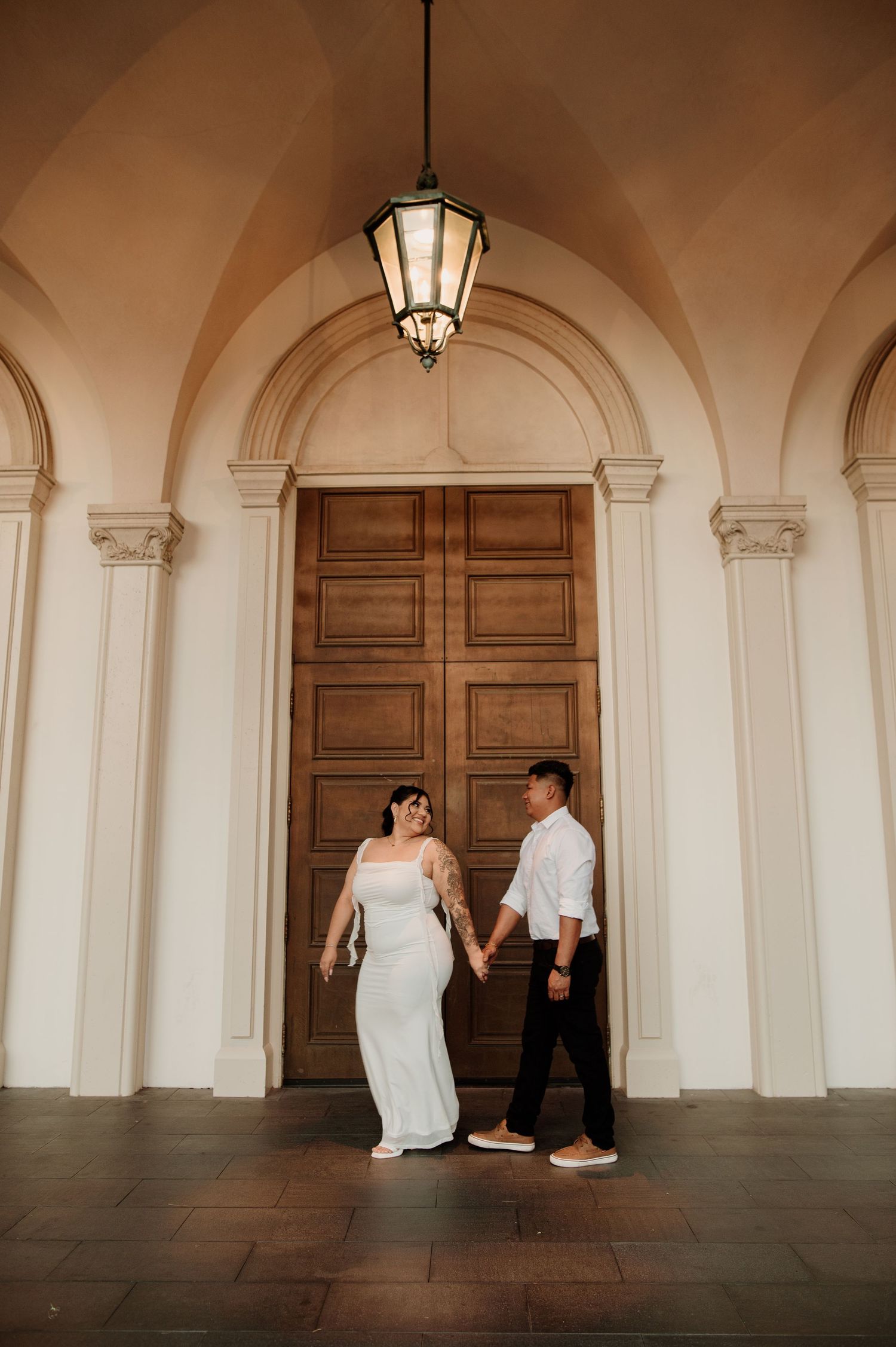 Bride in white gown and groom in casual attire hold hands beneath an elegant arched doorway with a vintage lantern.