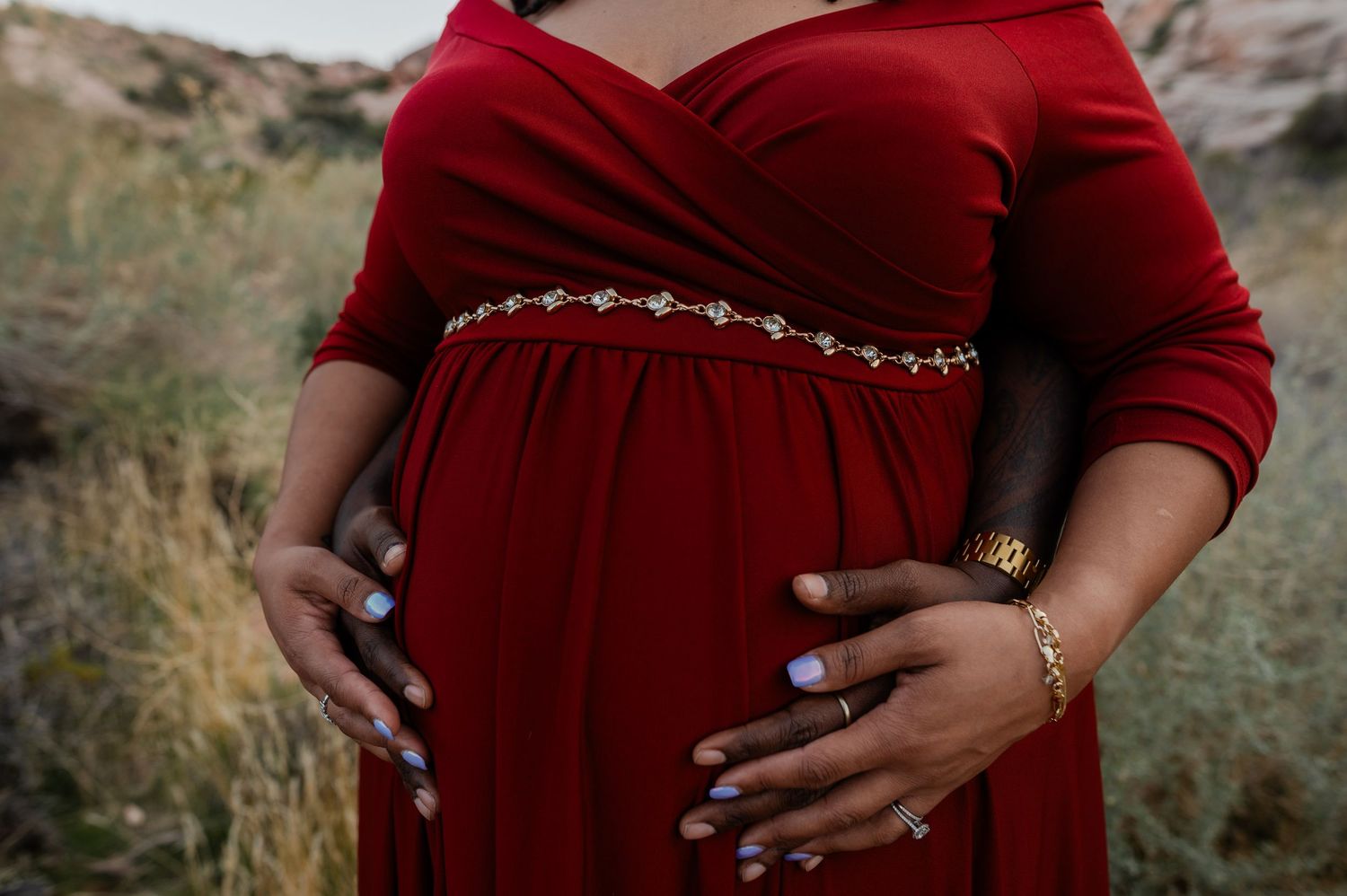 Close-up of pregnant belly in red maternity dress with jeweled belt and manicured hands.
