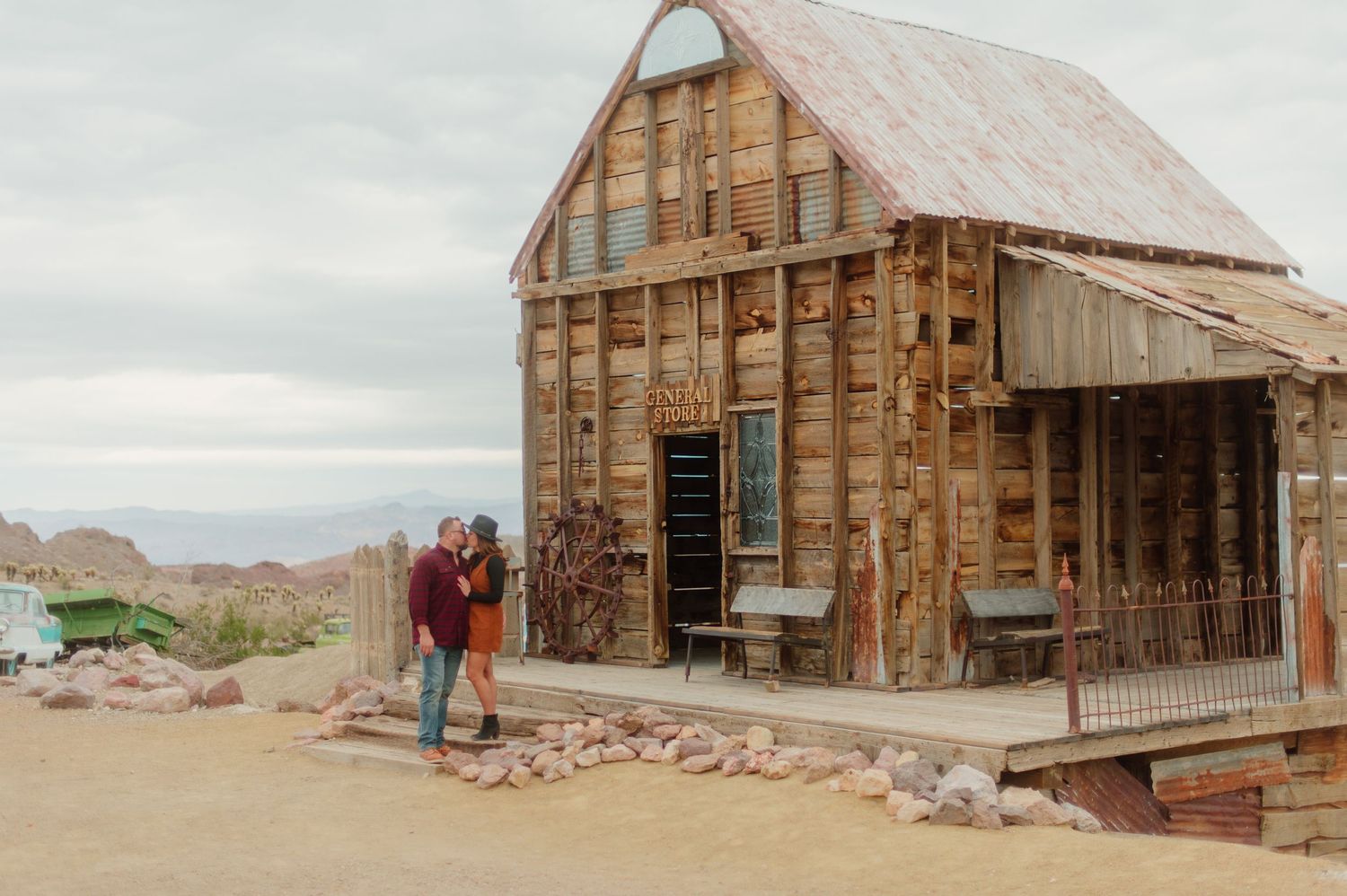 Couple embracing outside a rustic wooden building in a desert ghost town setting.