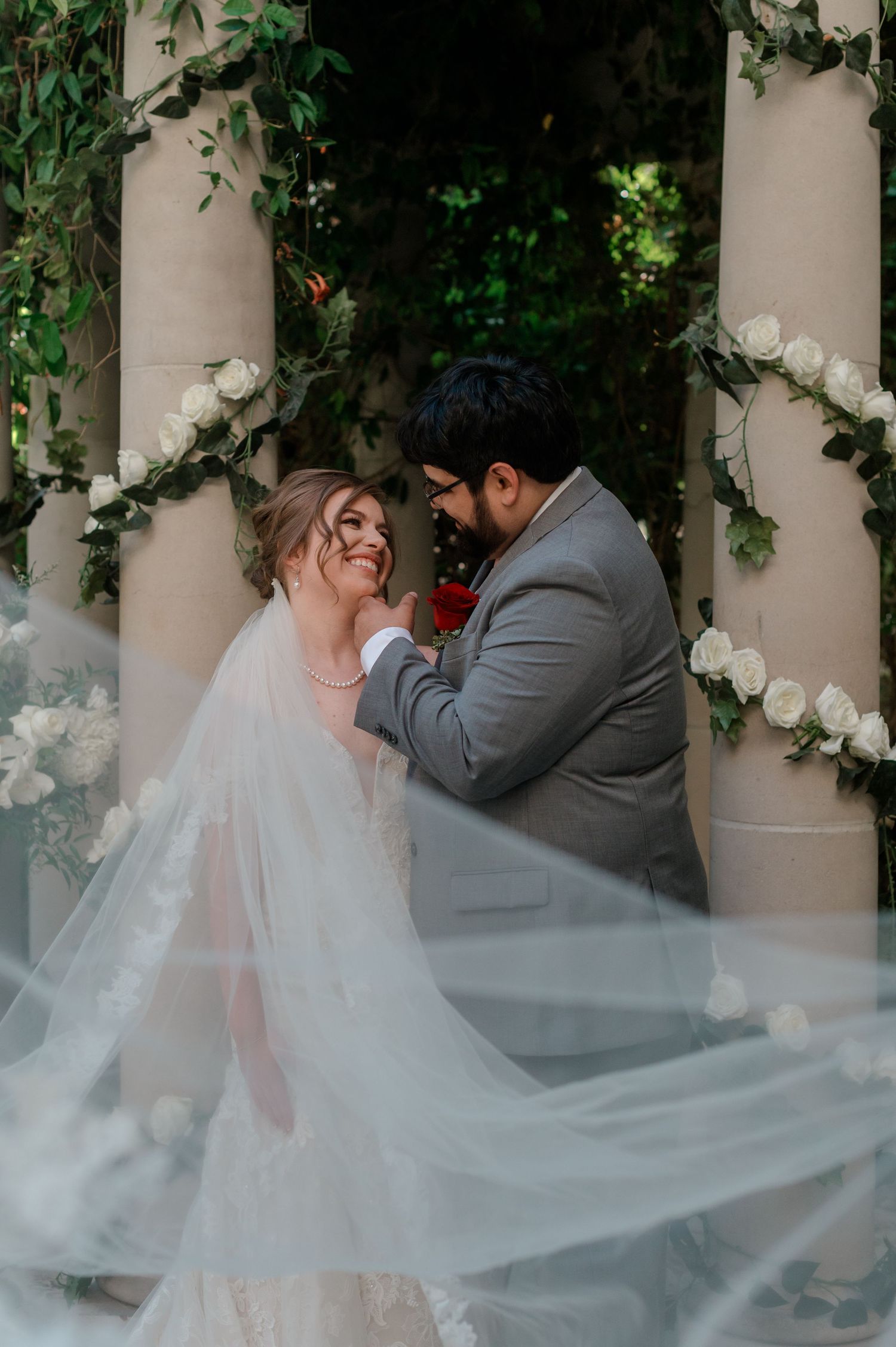 Bride and groom sharing a romantic moment under floral arch with white roses and greenery.