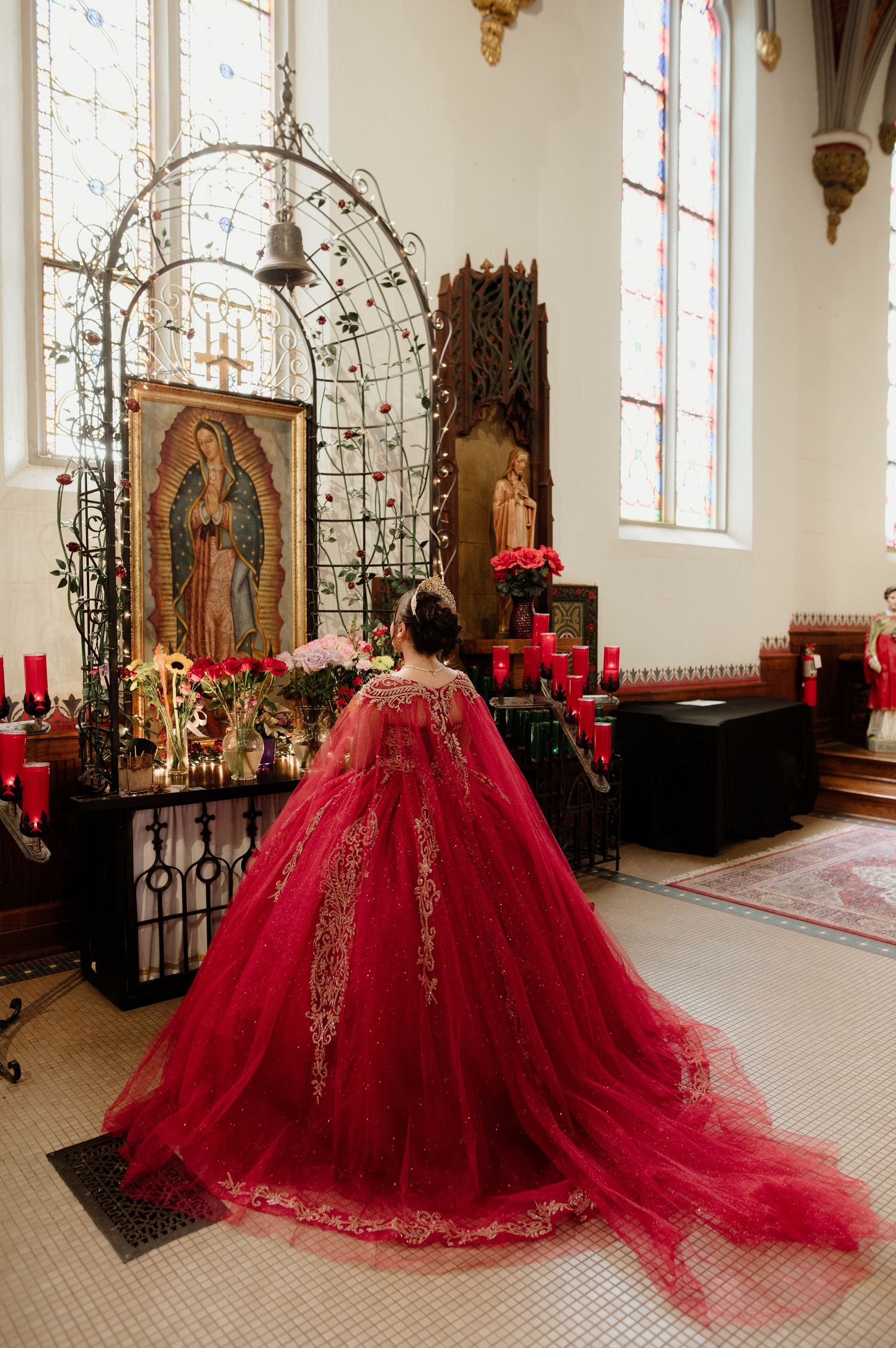 Quinceañera in red gown praying before Virgin of Guadalupe altar surrounded by red candles.