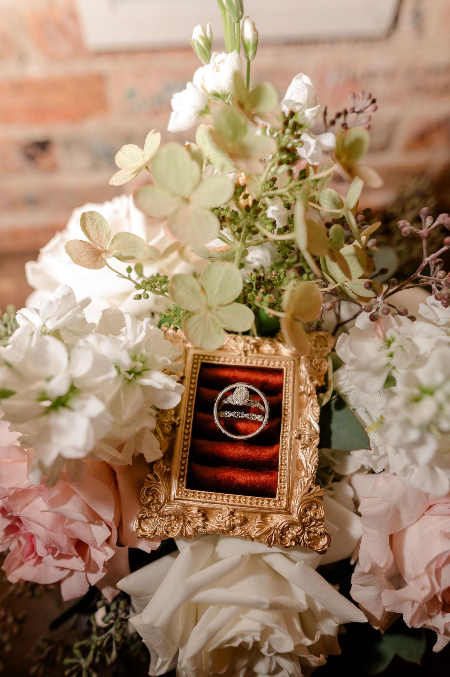 Wedding rings displayed in a gold ornate frame nestled among white and pink floral bouquet arrangements.