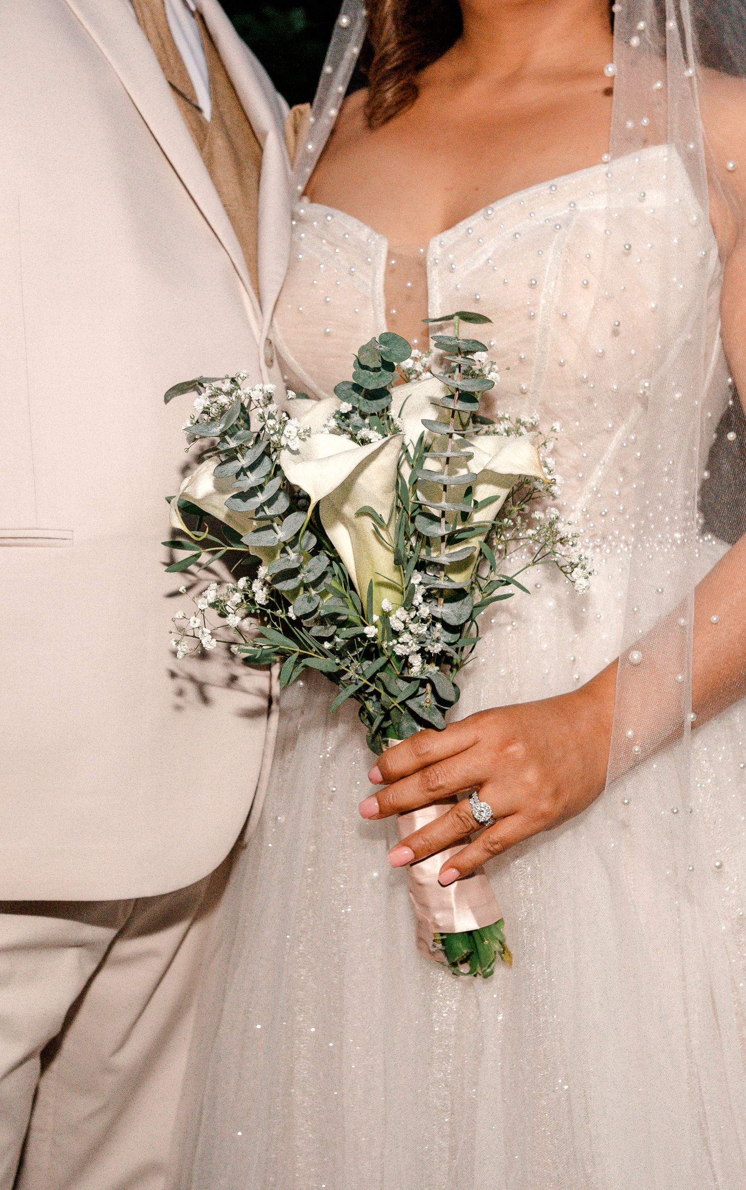 Bride holding white calla lily and eucalyptus bouquet in sparkly wedding gown beside groom in beige suit.