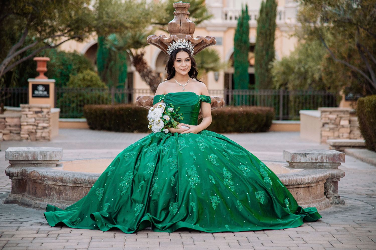 Quinceañera in a stunning emerald green embroidered ball gown and crown holding bouquet at an elegant outdoor fountain.
