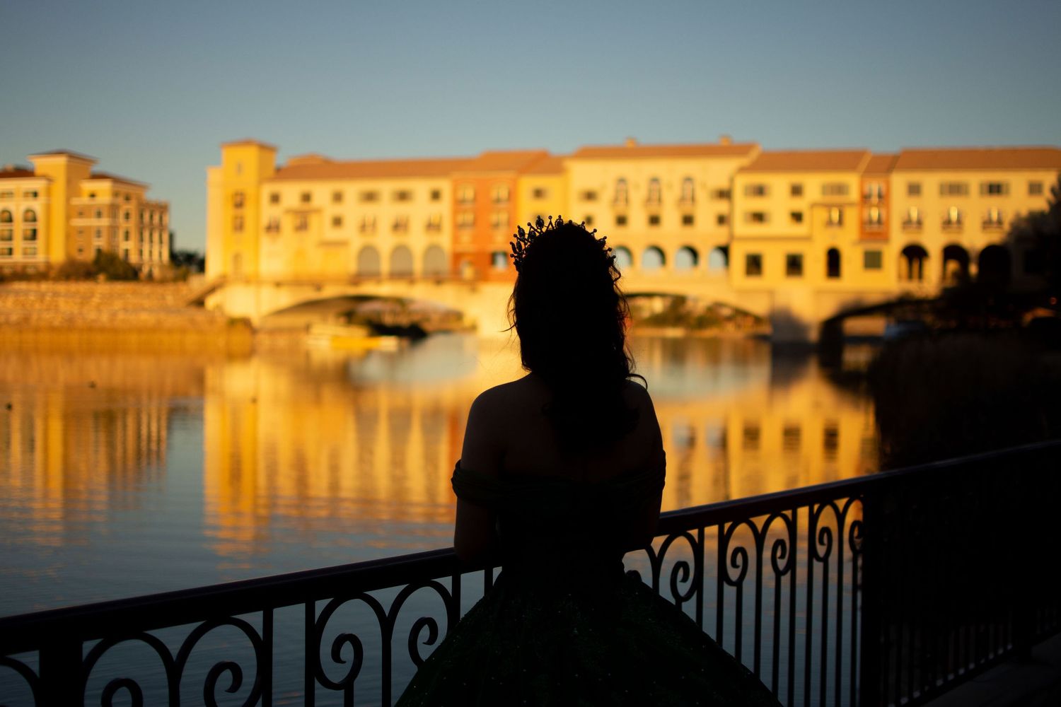 Silhouette of a quinceañera wearing a crown standing by a railing at golden hour with a lake and buildings behind.