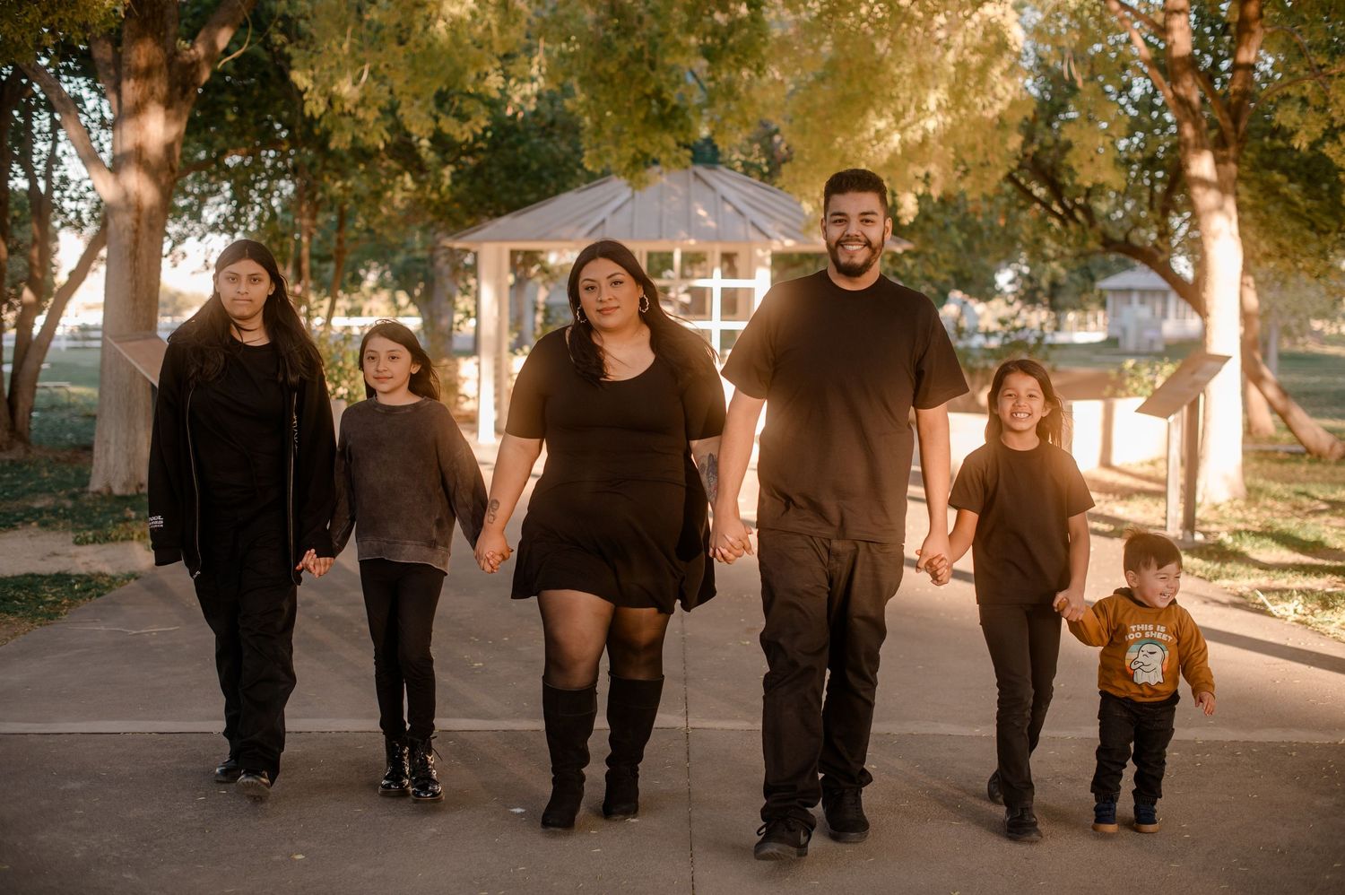 Family of six walks together through a tree-lined park in coordinating black outfits at golden hour.