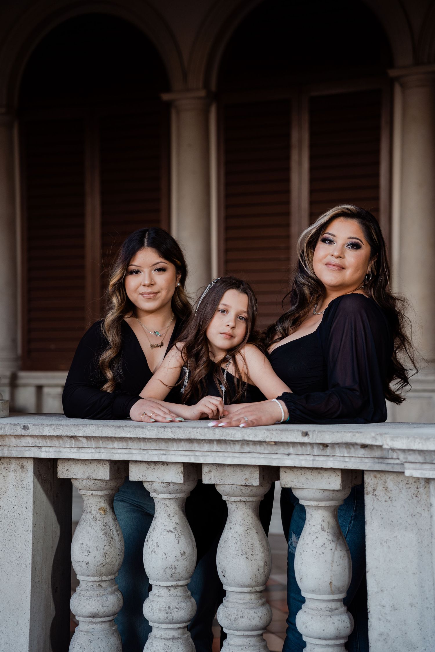 Three women in matching black outfits pose for a portrait on a decorative stone balcony railing.