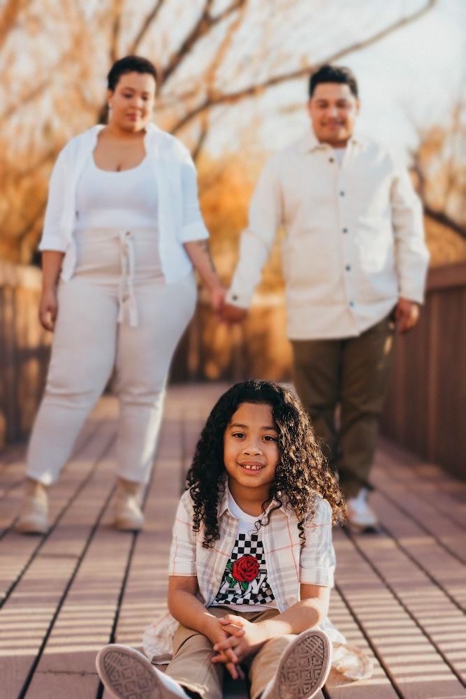 Smiling curly-haired child sits on wooden bridge while parents hold hands walking behind in autumn park.