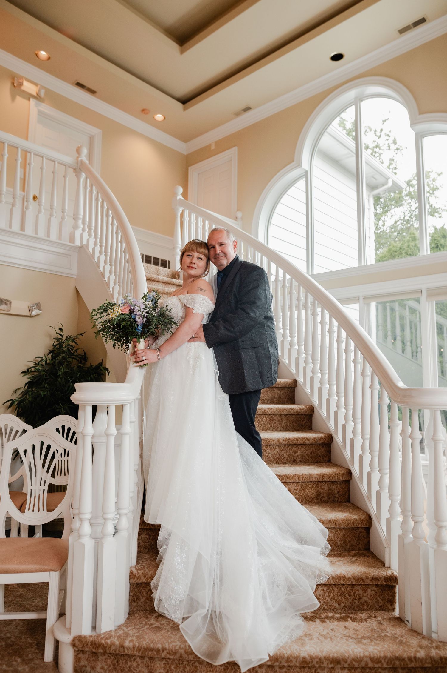 Bride and groom pose elegantly on a grand white staircase, bride holding a bouquet in a flowing white gown.