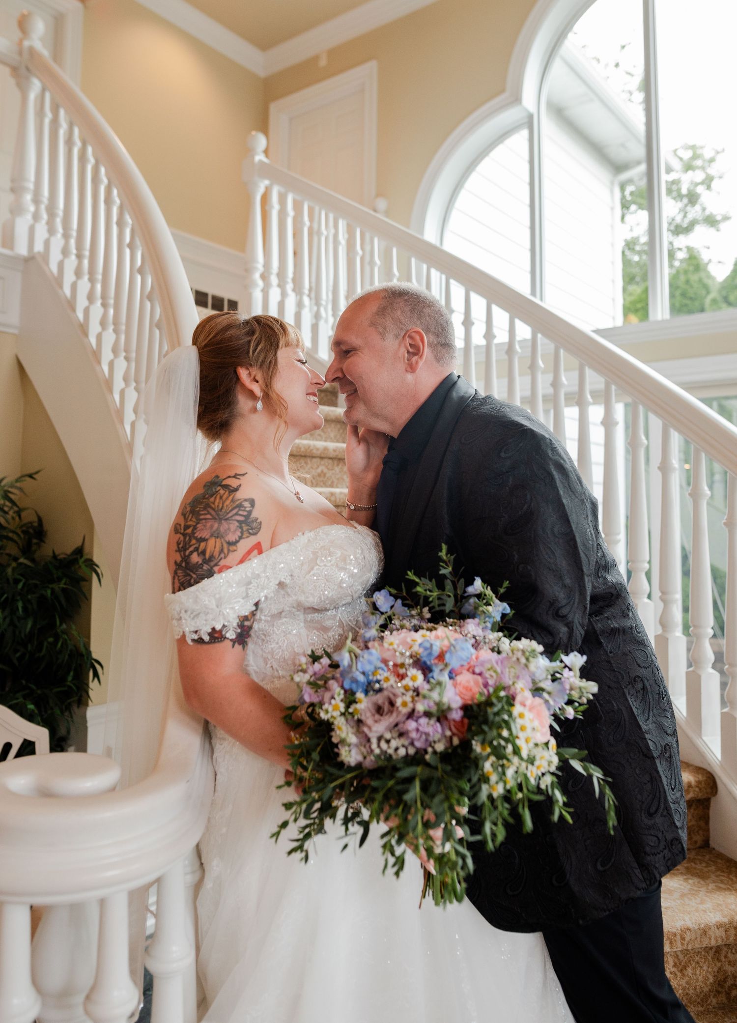Bride and groom share a romantic kiss on elegant white staircase, bride holding colorful wildflower bouquet in wedding venue.