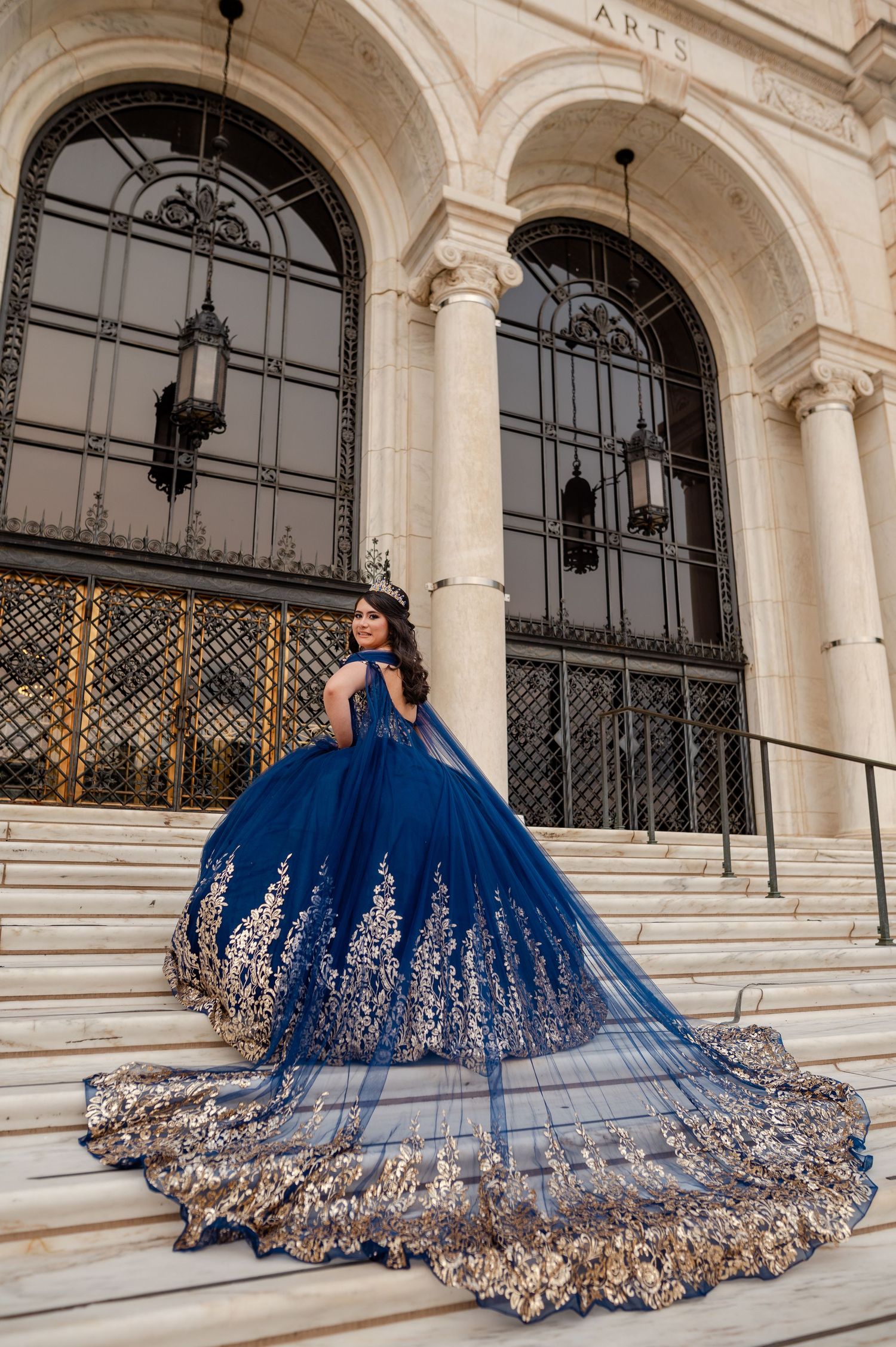 Young woman in navy blue and gold quinceañera ball gown poses on grand marble steps of elegant building.