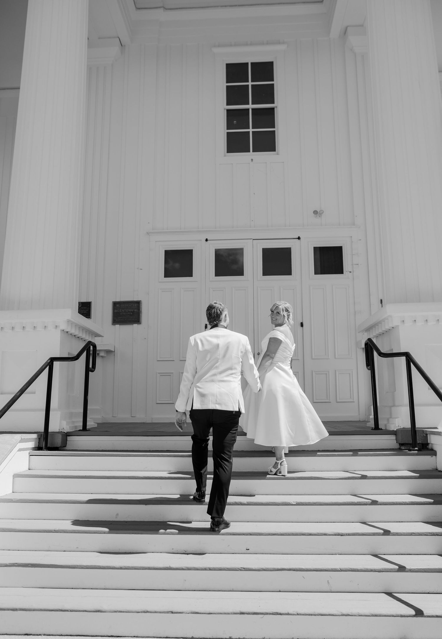 Black and white photo of bride and groom holding hands walking up church steps on wedding day.