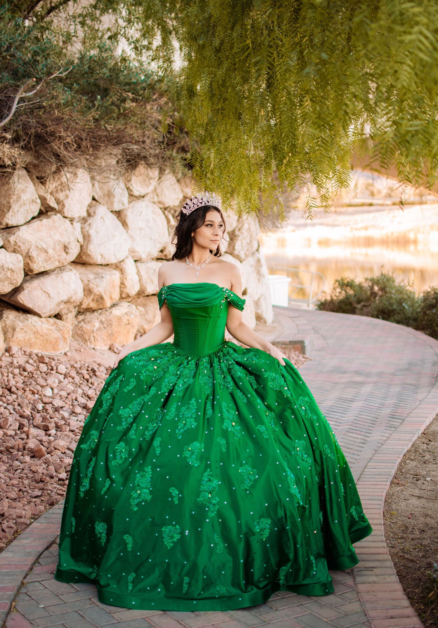 Young woman in a sparkling emerald green quinceañera gown walks along a brick garden path.